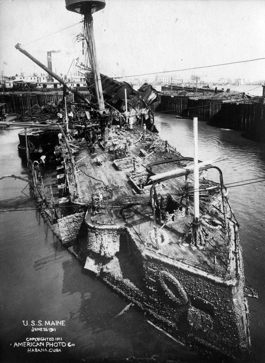 A historic photograph showing the wreck of USS Maine during salvage operations in Havana Harbor in 1911. The battered hull of the battleship sits inside a massive cofferdam, with pumps and machinery working to remove water. Wooden scaffolding and cranes surround the site, highlighting the scale of the engineering effort. Workers can be seen on and around the structure, tending to equipment and securing the wreck. This salvage allowed for the recovery of human remains and artifacts from the ship. It was one of the most complex naval recovery operations attempted at the time. The image documents the final chapter of the Maine’s story before she was towed out to sea and ceremonially sunk.