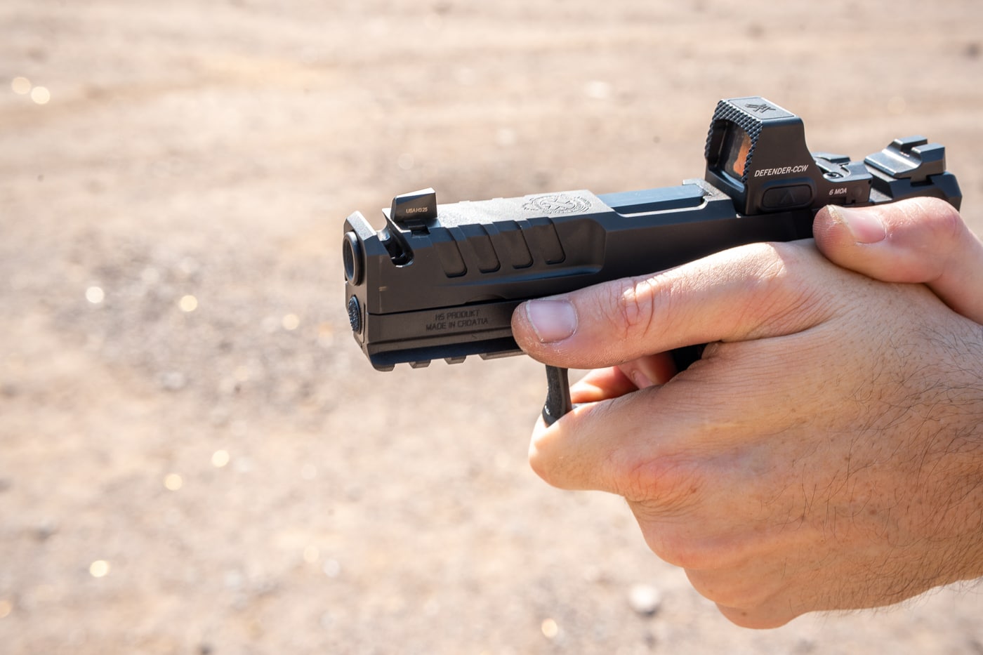 A close-up photograph of the Springfield Armory Echelon 4.0C Comp 9mm pistol being fired at an outdoor range, with the slide in mid-cycle and a spent casing ejecting. The image clearly shows the integral single-port compensator machined into the front of the slide and barrel, designed to vent gases upward to reduce muzzle rise. The pistol’s black Melonite finish is visible along with serrations and precision-machined details. A red dot optic is mounted directly to the slide, co-witnessing with the tritium U-Dot sights. The shooter’s hands are in a two-handed grip, demonstrating the pistol’s Adaptive Grip Texture. The background is blurred, drawing focus to the firearm’s features and recoil action. The image highlights both the engineering detail and live-fire performance of this compact, compensated handgun.