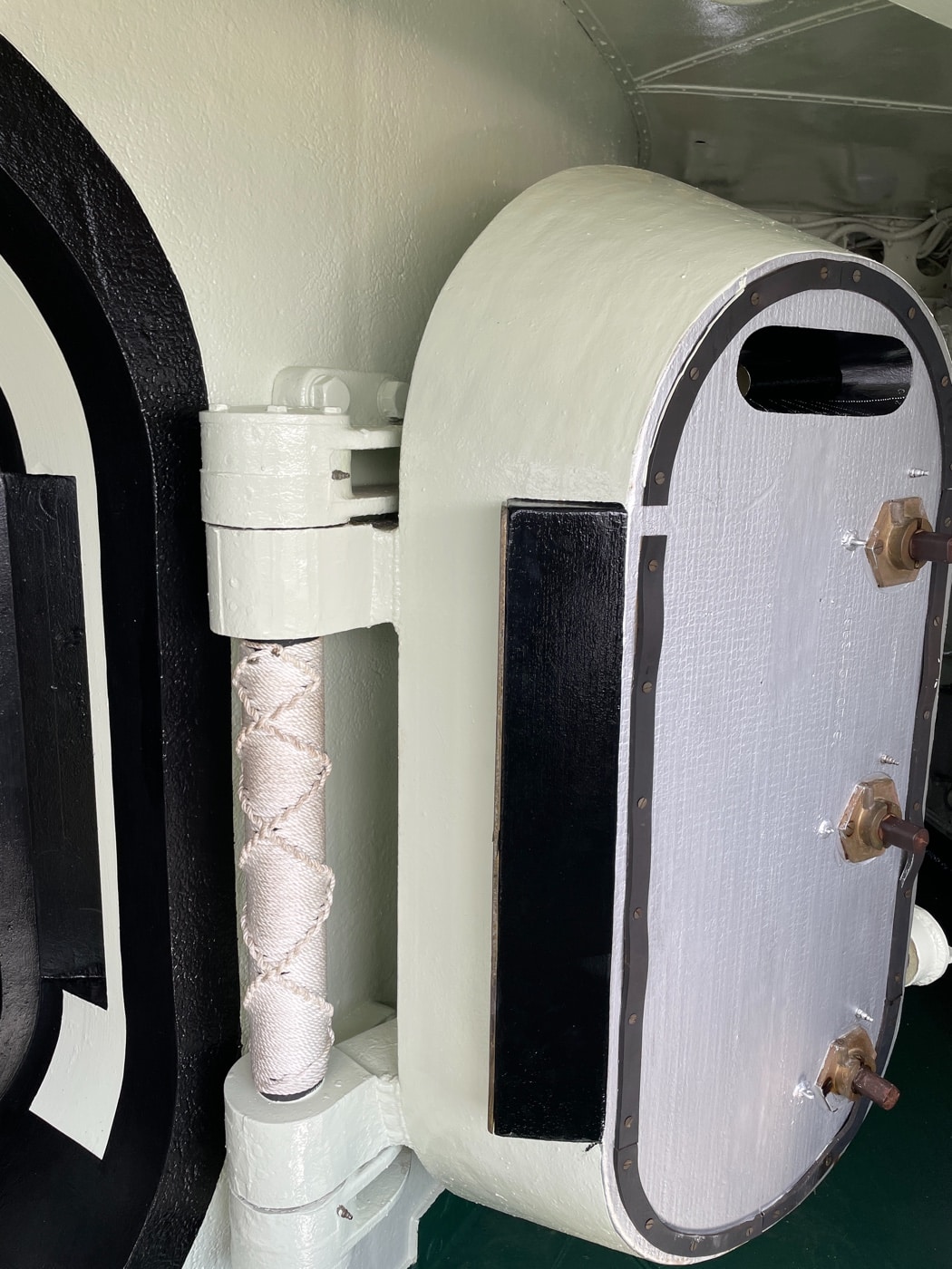 Close-up photo of the armored hatch leading into the USS Alabama’s bridge, highlighting the heavy construction and thickness of the steel door. The hatch is secured with robust hinges, locking mechanisms, and possibly a watertight seal. The surrounding bulkhead is also made of thick, reinforced armor plating, with visible rivets and welds. Lighting may show the texture and color of the steel, which is painted gray to match the rest of the ship. Any placards, warning signs, or operational instructions are visible nearby. The photo emphasizes the protection provided to the bridge and command personnel during ship-to-ship combat. This view helps illustrate the battleship’s focus on survivability and armored defense.