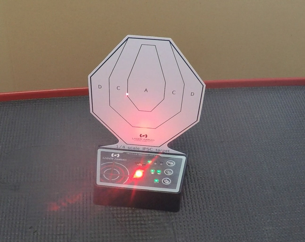 Close-up view of a Laser Ammo i-MTTS interactive target glowing bright red and producing an audible beep after detecting a laser hit. The target is mounted on its black electronic base and has a circular reflective face. This visual and audio feedback is part of the target’s training modes, helping shooters track accuracy without live fire. The system works with AR15 rifles equipped with the Flash Bolt as well as handguns using laser cartridges. The red illumination indicates a confirmed hit, reinforcing proper trigger control and aiming technique. The scene is set indoors, demonstrating safe practice in a controlled environment.