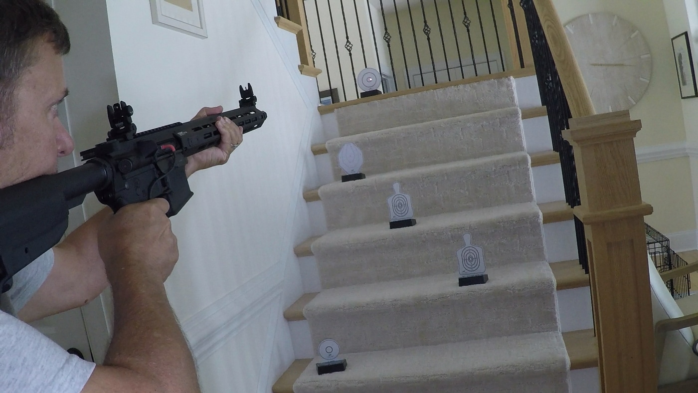 Several Laser Ammo i-MTTS interactive targets placed on the steps of a residential staircase for dry-fire training. The targets are mounted on their electronic bases and feature reflective surfaces shaped as bullseyes and silhouettes. Each unit runs on AAA batteries and can be set to different training modes. The spacing between steps creates varied engagement angles for the shooter. The setup is designed for use with Laser Ammo’s Flash Bolt or laser cartridges. The environment is a typical home setting, emphasizing safe, no-ammo training indoors.