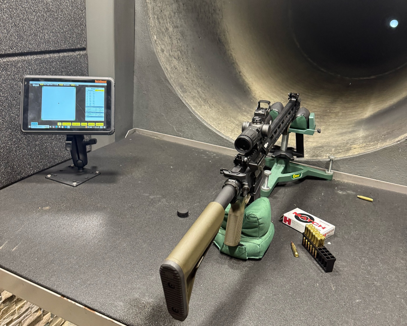The image shows an indoor rifle shooting range where the author is testing the EOTECH Vudu 3-9X32 riflescope mounted on a Springfield Armory SAINT AR-15. The shooter is positioned in a booth with a bench rest, aiming at a paper target down a long, dimly lit range. Hearing protection and safety glasses are worn for safety. The rifle is OD Green, and the black Vudu scope is mounted with an integrated base. The target is visible far in the background, with overhead lighting casting a warm glow on the shooting lane. This setup is used to confirm zero and evaluate scope clarity under indoor lighting.