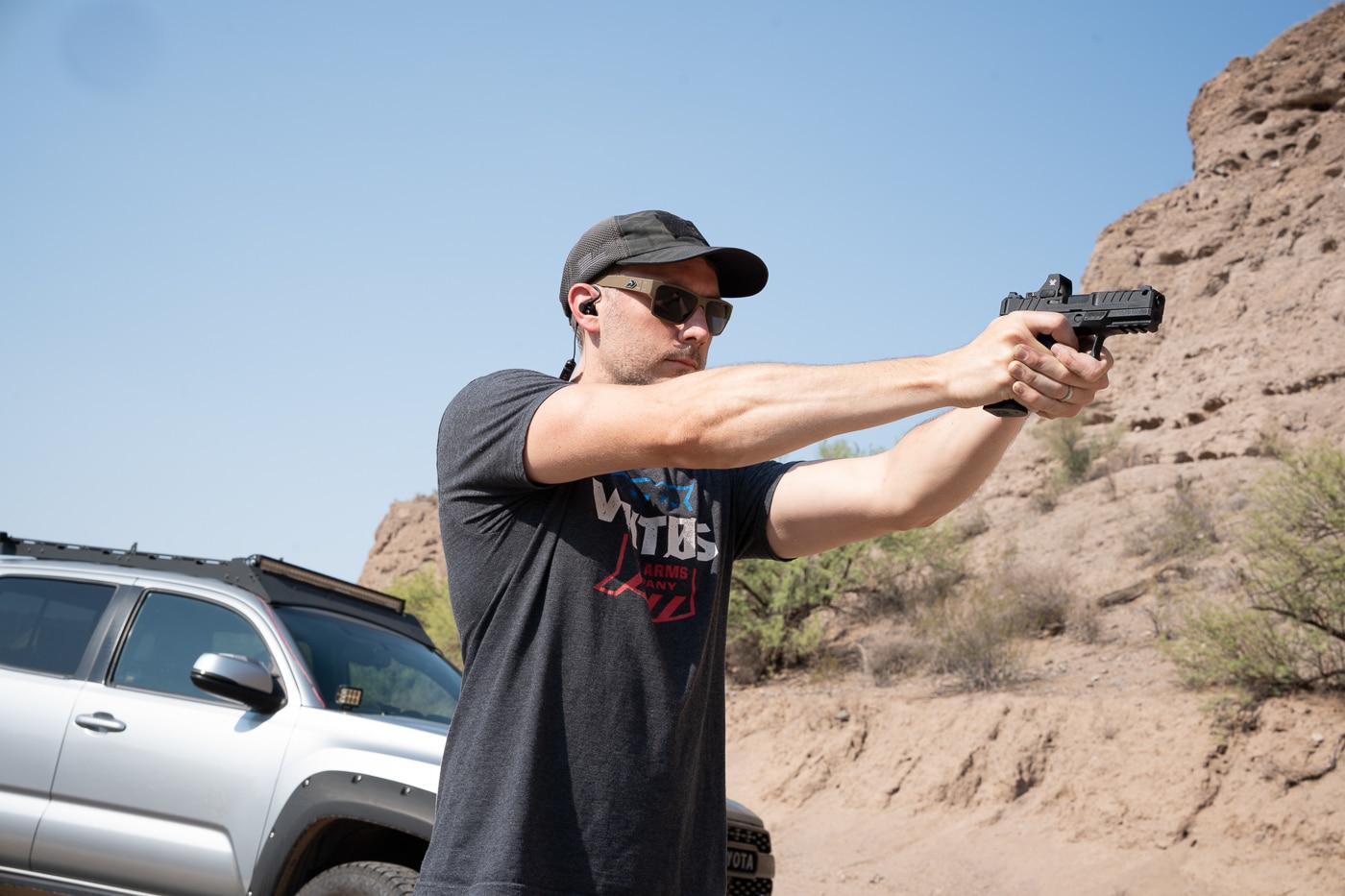 A low-angle photograph captures the author firing the Springfield Armory Echelon 4.0C Comp 9mm pistol at an outdoor range. The image emphasizes the front of the handgun, with the integral single-port compensator visible just behind the tritium/luminescent front sight. The slide is in motion, ejecting a spent casing, and the black Melonite finish reflects the natural lighting. A red dot optic is mounted directly to the slide, co-witnessing with the U-Dot rear sight. The shooter maintains a firm two-handed grip, displaying the Adaptive Grip Texture. The upward venting of gases from the compensator can be seen, illustrating its role in reducing muzzle rise. The low viewpoint gives prominence to the pistol’s design and live-fire action.