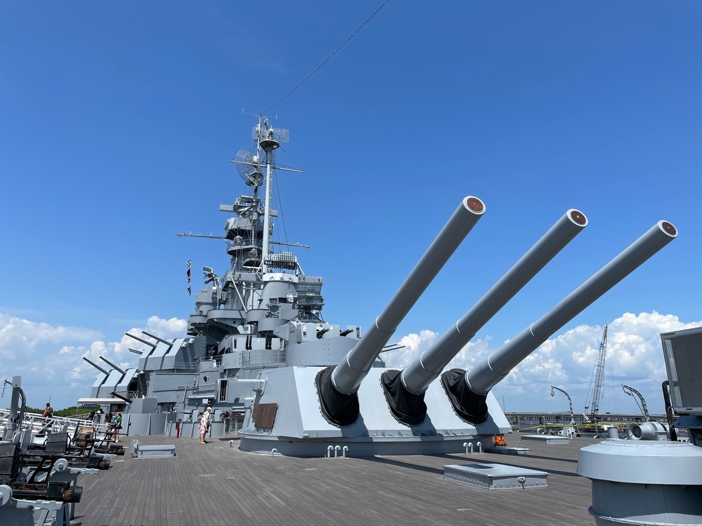 Photo of USS Alabama’s main gun turrets, featuring a total of nine 16-inch rifled cannons arranged in three triple turrets. The shot focuses on the forward section of the battleship, with the gun barrels pointing out to sea or raised at various angles. The steel turrets are heavily armored, with mechanical details and access hatches visible on their surfaces. The scale of the guns is emphasized by comparing them to the surrounding deck fittings, railings, and any crew members present. The sky is clear, highlighting the powerful silhouette of the ship’s primary weapons. This image illustrates the main offensive capability of the South Dakota-class battleship. The scene underscores the historical significance and engineering of these naval guns.