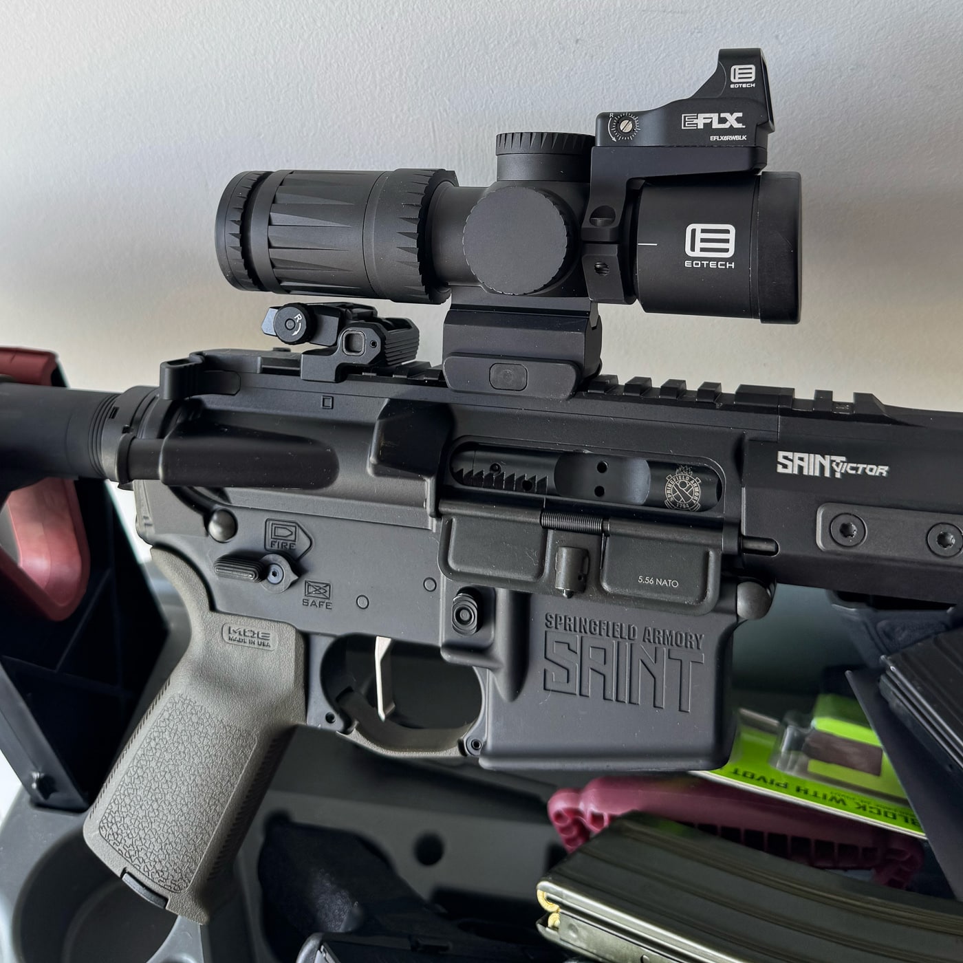 The image shows a Springfield Armory SAINT Victor AR-15 rifle with an EOTECH Vudu 3-9X32 second focal plane riflescope mounted to the top Picatinny rail. The scope is black with capped turrets and an integrated base mount, giving it a clean and streamlined look. The rifle’s OD Green finish contrasts with the optic’s darker color. A small red dot sight is mounted above the scope for close-quarters use. The rifle rests on a shooting bench, with bright outdoor lighting making the details of the rifle’s receiver, barrel, and optic clear. The compact size of the Vudu scope stands out against the full-length rifle platform.