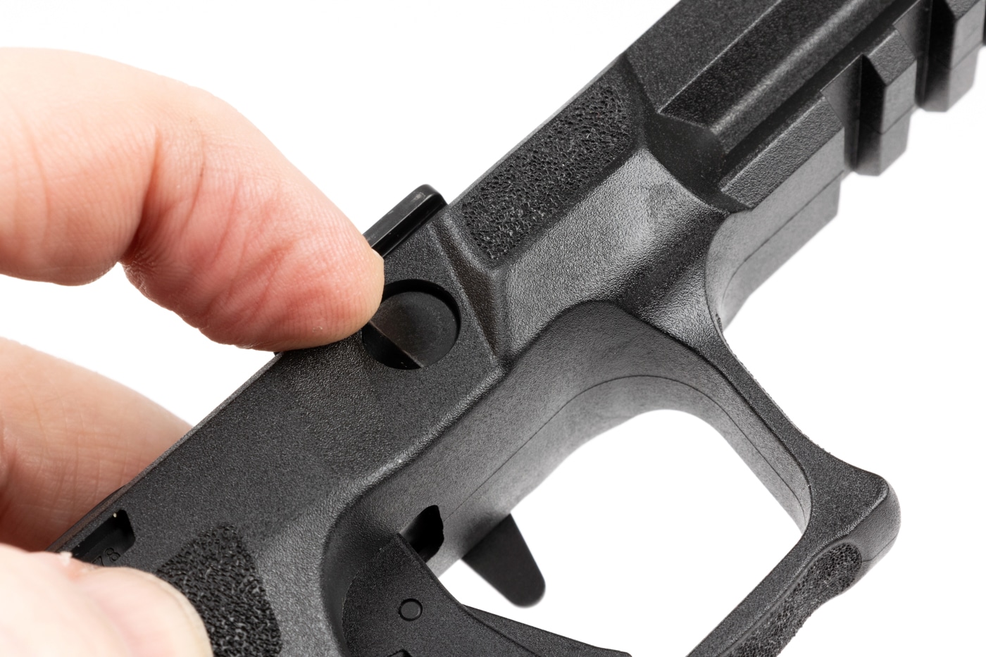 The image shows a close-up of a person using their finger to push in the flat end of the disassembly lever on the right side of a Springfield Echelon 4.0C pistol. The gun is partially disassembled, with the slide removed, and the focus is on the disassembly lever just above the trigger area. The person’s hand is positioned to apply gentle pressure, as recommended in the user manual. The background is neutral and uncluttered, making it easy to see the location and movement of the lever. This step is necessary to free the disassembly lever so it can be pulled from the left side of the grip module. The photo highlights that no special tools are needed — just a finger or non-marring tool. It’s a clear demonstration of the first step in removing the Central Operating Group for a grip swap.