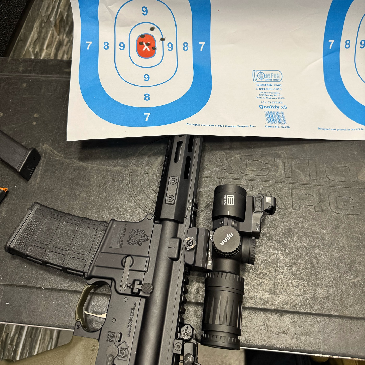 The image shows a Springfield Armory SAINT AR-15 with an EOTECH Vudu 3-9X32 riflescope mounted on its top rail, positioned next to a paper target. The target features a tight grouping of bullet holes clustered near the bullseye, demonstrating the optic’s precision. The rifle has an OD Green finish, while the compact black Vudu scope is clearly visible. The photo is taken in bright daylight, with the rifle resting on a shooting bench. The grouping indicates the rifle and optic were properly zeroed and capable of consistent accuracy.