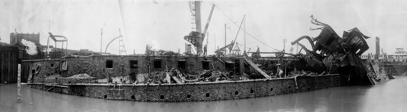 A historic photograph of USS Maine during salvage operations in June 1911, showing the ship partially raised inside a cofferdam in Havana Harbor. The hull is exposed above the waterline, with the bow end heavily damaged and bent from the 1898 explosion. Large cracks, twisted plating, and missing sections reveal the force of the blast that destroyed the ship. Wooden supports, scaffolding, and machinery surround the wreck as engineers worked to stabilize it. Workers can be seen along the structure, underscoring the scale of the operation. The image documents one of the most significant stages of the salvage, when the extent of the destruction became visible for the first time. This view highlights both the tragedy of the Maine and the complexity of early 20th-century naval recovery efforts.