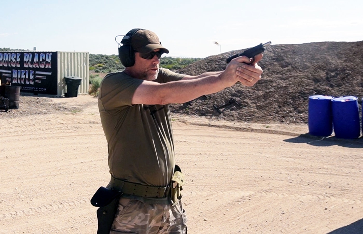 The photo shows the author actively shooting the Springfield Armory Mil-Spec 1911 pistol at an outdoor shooting range. The author is in a typical shooting stance, arms extended, holding the parkerized steel 1911 with both hands. Spent brass may be visible in the air, indicating the pistol has just been fired. Range targets and safety barriers are present in the background, with typical outdoor lighting and a safe, open environment. The author is wearing appropriate eye and ear protection. This image visually demonstrates the Mil-Spec 1911’s use in a real range setting, helping to illustrate the article’s points about the gun’s performance and upgrades over the original G.I. models. The photo is ideal for content about shooting the Springfield Mil-Spec 1911, range testing, or evaluating modern 1911 handguns.