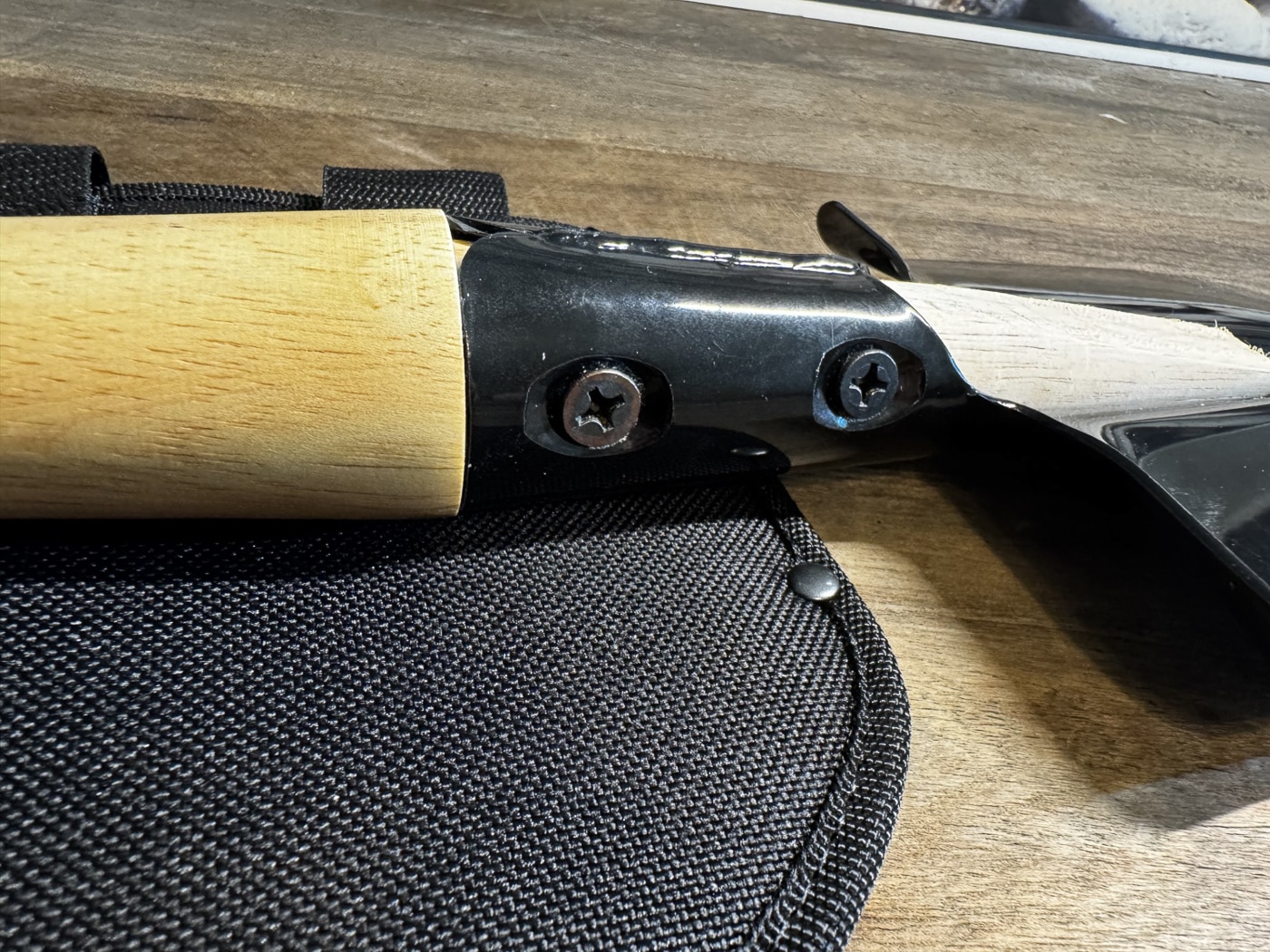 Close-up photograph of the Cold Steel Special Forces Shovel where the hickory handle meets the steel blade socket. The steel neck is fully welded along the seam for reinforcement. Two large Phillips head screws secure the handle into the socket, providing a tight, stable fit. The steel surface has a matte black protective coating, while the wood shows natural grain and lathe marks from manufacturing. The image emphasizes the durability of this attachment method. This joint design minimizes flex under digging, chopping, or prying forces.