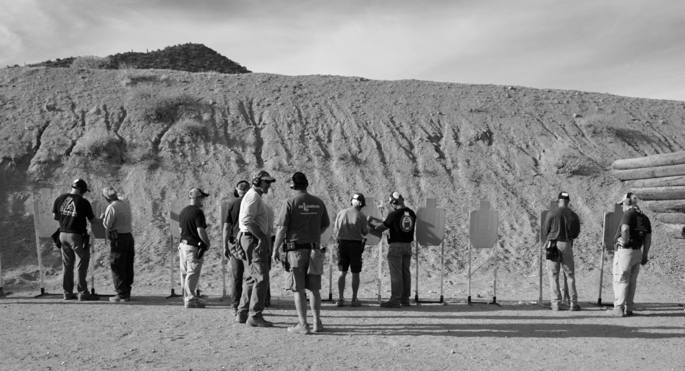 Several students stand side by side on a shooting range, each aiming their pistols downrange as an instructor oversees their technique. The scene shows various stages of shooting, with participants focused on their targets and using proper stance and grip. The instructor is present to correct form and offer advice, emphasizing the importance of professional instruction in developing hard skills. The range is outdoors, with target stands visible in the distance. This image illustrates the training process required to improve reaction speed and stay ahead of the power curve in a gunfight. It highlights the role of guided practice and repetition in building proficiency with firearms. Ideal for topics on shooting instruction, personal defense, and skill development.