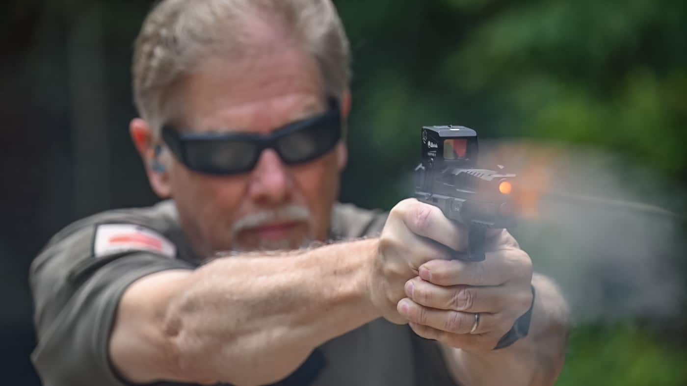 Photo shows the author in the act of shooting a Springfield Armory Echelon pistol fitted with a Burris FastFire E enclosed red dot sight. The author’s grip and stance are stable, both hands on the pistol, as a spent casing is ejected or smoke is visible at the muzzle. The FastFire E’s compact, enclosed housing is mounted securely on the slide, and the sight window is prominent. The shooting range background includes standard features like targets and shooting bays, with the author focused on aiming at a distant target. The image demonstrates the optic’s practical use and stability under recoil, emphasizing real-world evaluation. Lighting is balanced to highlight both the firearm and the mounted optic. This photo is suitable for illustrating the Burris FastFire E’s performance during a hands-on review.