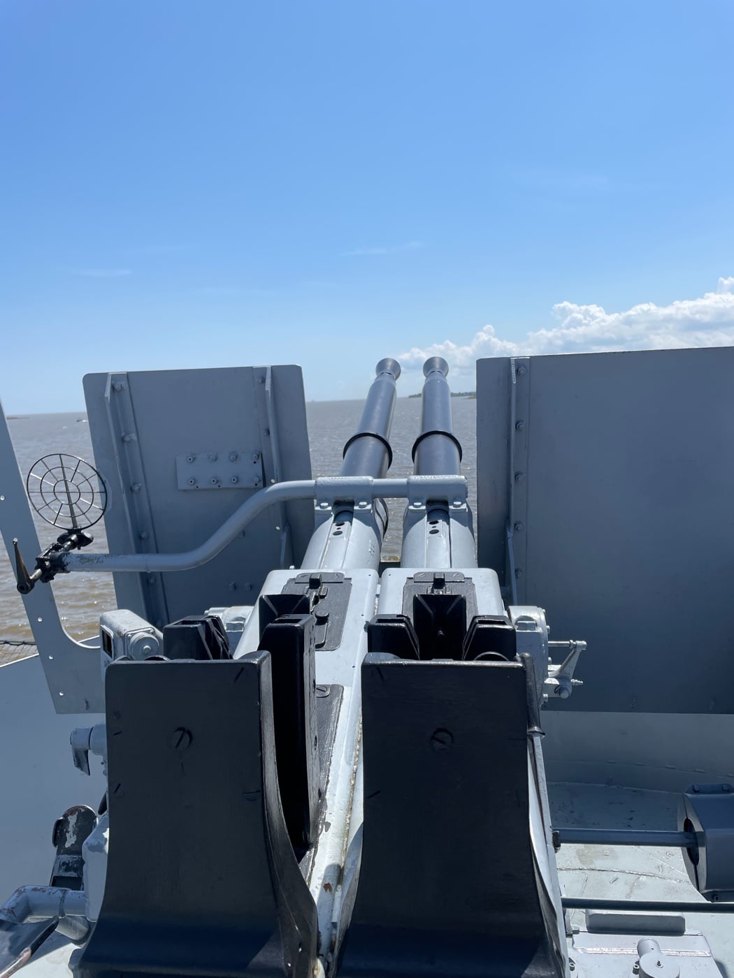 Photo showing a close-up of a twin mount 40mm Bofors antiaircraft gun installed on the deck of USS Alabama. The two gun barrels are positioned side by side within an open, armored mounting, with control wheels, gun sights, and protective shields visible. The mount is placed near the edge of the deck, with the ship’s railing, ocean, or other equipment in the background. Ammunition boxes or belt feeds can be seen nearby, indicating readiness for sustained firing. The structure is painted naval gray, consistent with World War II-era warships. Crew seating and operating controls are detailed, though no personnel are present in the shot. This image highlights the key role of the 40mm Bofors in defending the battleship from attacking aircraft. The scene is informative for understanding the ship’s layered air defense system.