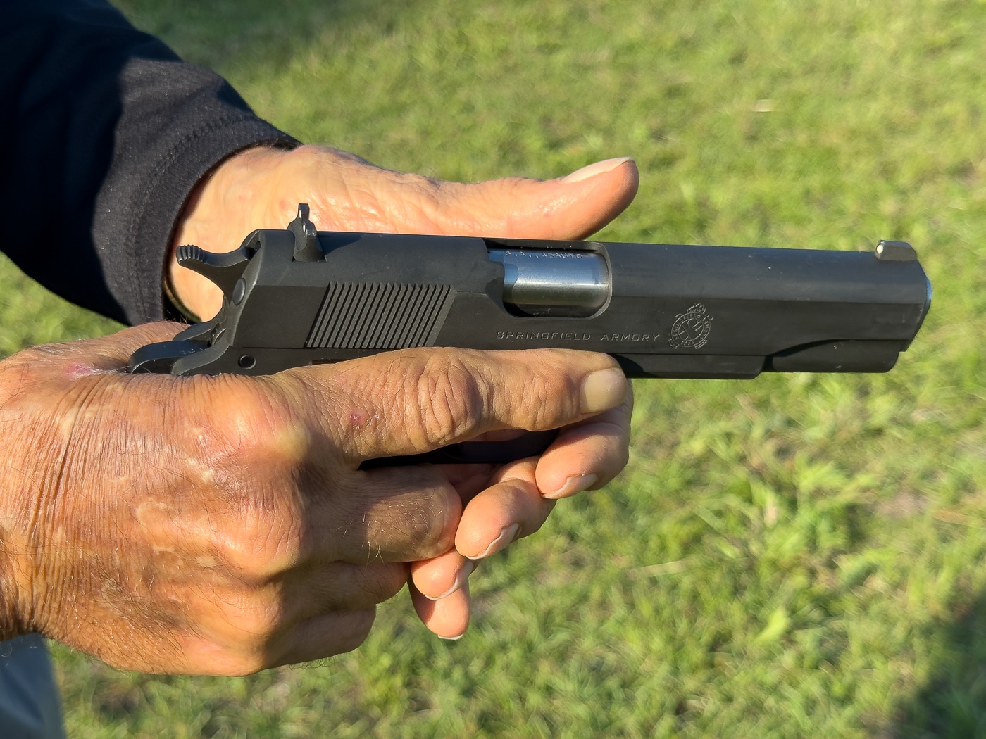 In this photo, the reassembly process of a 1911 pistol with a G.I. bushing is shown in detail. The slide is drawn back so that the slide stop can be aligned with the correct notch. A screwdriver tip presses the plunger rearward, holding it in place. The spring is left hanging out under the barrel bushing, making it easier to manage the components. The image highlights hand placement, the slide position, and the plunger being controlled. This step-by-step view demonstrates the safest way to insert the slide stop. It’s a useful instructional photo for anyone maintaining a 1911.