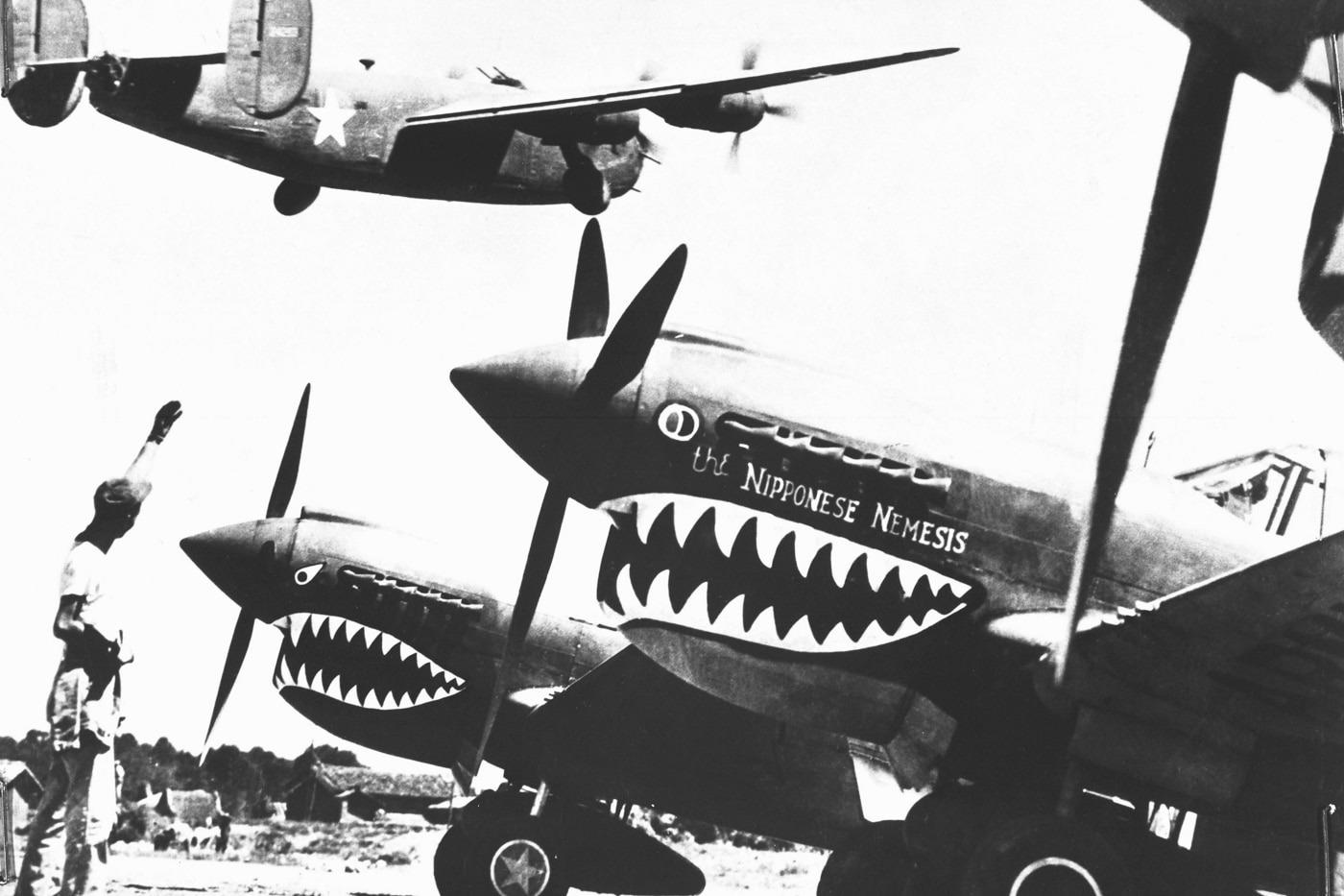 A Consolidated B-24 Liberator bomber is shown taking off from an advanced U.S. base in China in 1943, its landing wheels retracting into the fuselage. In the foreground, shark-nosed Curtiss P-40 fighter planes are parked, instantly recognizable by their iconic paint scheme. An American soldier stands nearby, waving to the departing crew as a gesture of good luck. The Liberator was tasked with long-range bombing missions against Japanese targets, using forward bases in China as staging points. This photo illustrates the cooperation between bomber and fighter units in the China-Burma-India theater. The rugged conditions of the base, visible in the dirt runway and dispersed aircraft, reflect the difficult environment of these operations. It captures both the humanity of the sendoff and the scale of the mission ahead.