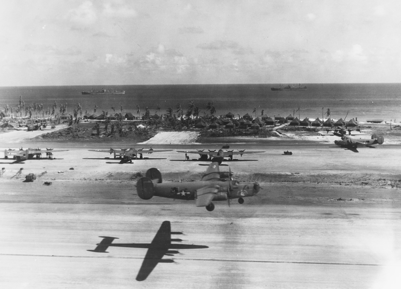 A U.S. Army Air Forces Consolidated B-24 Liberator bomber takes off from Eniwetok Island airfield in the Marshall Islands on April 13, 1944. The photograph, taken from the top of an observation tower, shows the bomber climbing from the coral-surfaced runway. In the distance, several other B-24s are parked along the airfield, prepared for upcoming missions. Beyond the shoreline, two Liberty ships are anchored offshore, providing logistical support to the base. The scene illustrates how remote Pacific atolls were transformed into strategic airfields capable of launching long-range strikes. Eniwetok was an important staging point for raids on Japanese-held islands. The photo combines aviation, naval supply, and base construction in one moment of wartime activity.