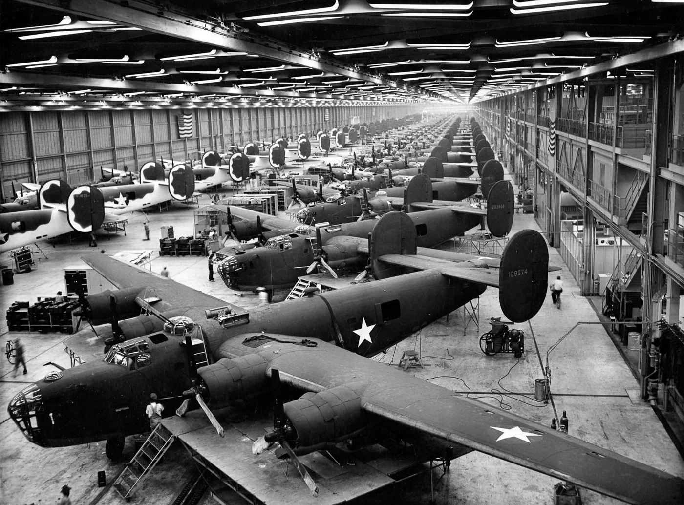 A wide view of the Consolidated B-24 Liberator heavy bomber assembly line during World War II, showing multiple aircraft in various stages of construction. Rows of partially completed fuselages stretch across the factory floor, with workers using scaffolds and platforms to access different sections. The factory space is filled with cranes, workbenches, and production equipment supporting the massive industrial effort. The image highlights the scale of American wartime manufacturing, with dozens of bombers being built at once. The B-24 was produced in record numbers, with over 18,000 completed by the war’s end. This photo reflects the speed and efficiency of the assembly line process, a key factor in Allied air power. The scene captures the transformation of raw materials into one of history’s most important bombers.