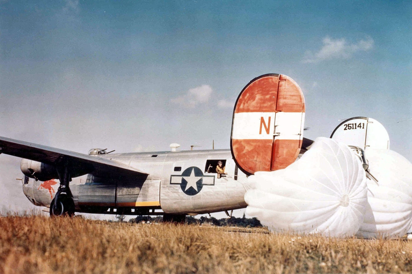 A Consolidated B-24H Liberator bomber is shown landing at RAF Woodbridge airfield in Norfolk, England, with drag parachutes deployed behind the aircraft. The parachutes billow open, helping slow the heavy four-engine bomber as it touches down on the long runway. RAF Woodbridge, constructed during World War II, was specifically designed as an emergency landing field for damaged Allied bombers returning from missions over Europe. The photo captures the size and bulk of the Liberator as well as the challenges of safely landing after long combat sorties. The deployed parachutes reduced strain on the brakes and shortened landing distance. This image highlights the engineering measures used to handle the heavy, high-speed landings of wartime bombers. It also illustrates the importance of specialized airfields like Woodbridge to the survival of many bomber crews.