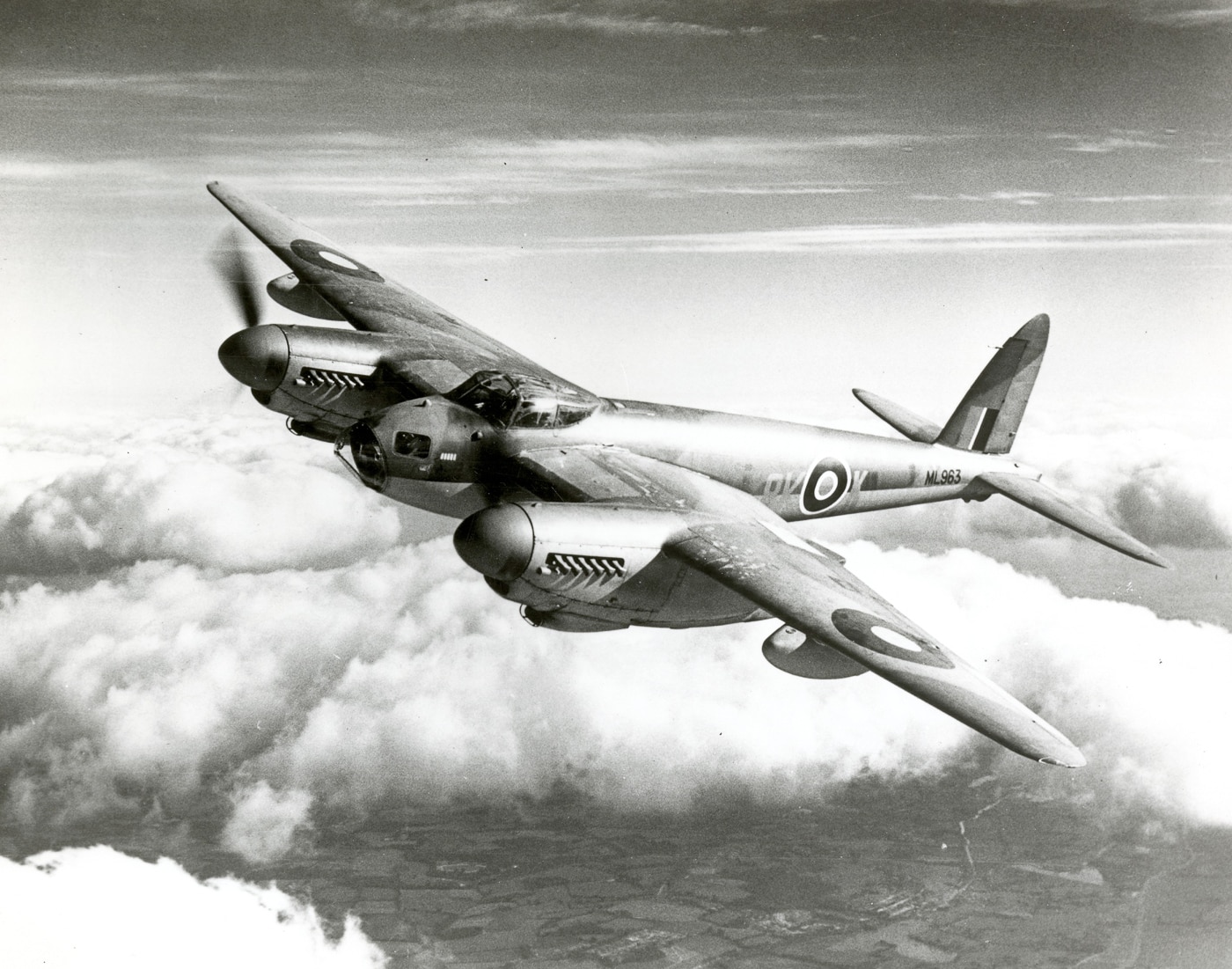 A de Havilland Mosquito aircraft captured mid-flight against a clear sky, showing its sleek wooden construction and twin Rolls-Royce Merlin engines. The Mosquito was known for its speed, often outrunning German fighters during World War II. Its lightweight balsa and plywood airframe allowed for rapid production and reduced reliance on scarce metals like aluminum. This particular Mosquito is painted in authentic wartime camouflage colors. The aircraft was used in a wide range of missions including precision bombing, reconnaissance, and night fighting. Its adaptability made it one of the most effective British aircraft of the war. The image highlights the narrow fuselage and broad wings that gave the Mosquito its exceptional performance.