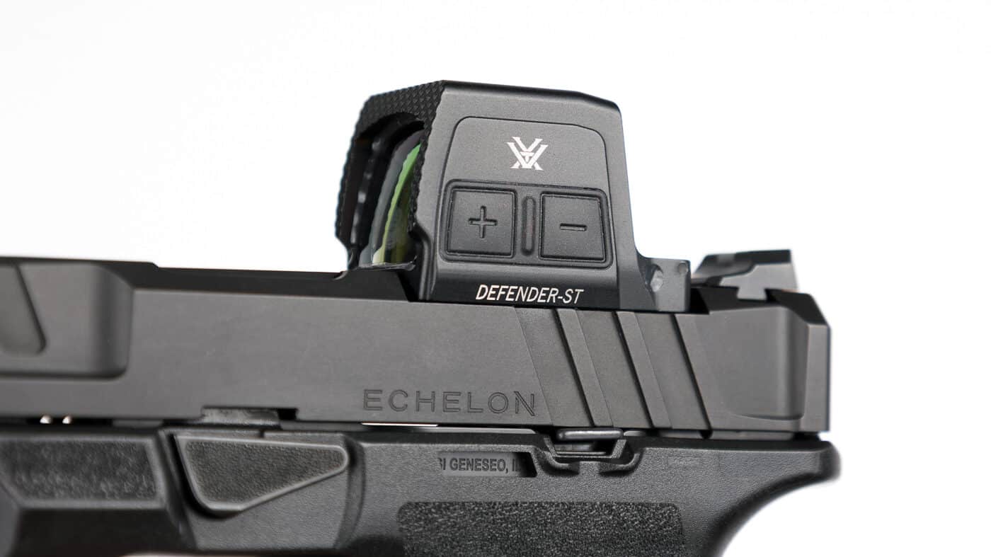 Close-up view of the Vortex Defender-ST Enclosed red dot sight showing the side-mounted brightness controls. Two rubberized buttons, marked with plus and minus symbols, are positioned on the left side of the optic housing. These buttons adjust reticle brightness across twelve levels, including two that are compatible with night-vision devices. Holding both buttons simultaneously cycles through the optic’s three reticle options: 3 MOA dot, 32 MOA circle, or circle-dot combination. The controls are recessed slightly into the black anodized aluminum housing to prevent accidental activation during concealed carry or duty use. The image highlights how accessible and intuitive the controls are without adding bulk.