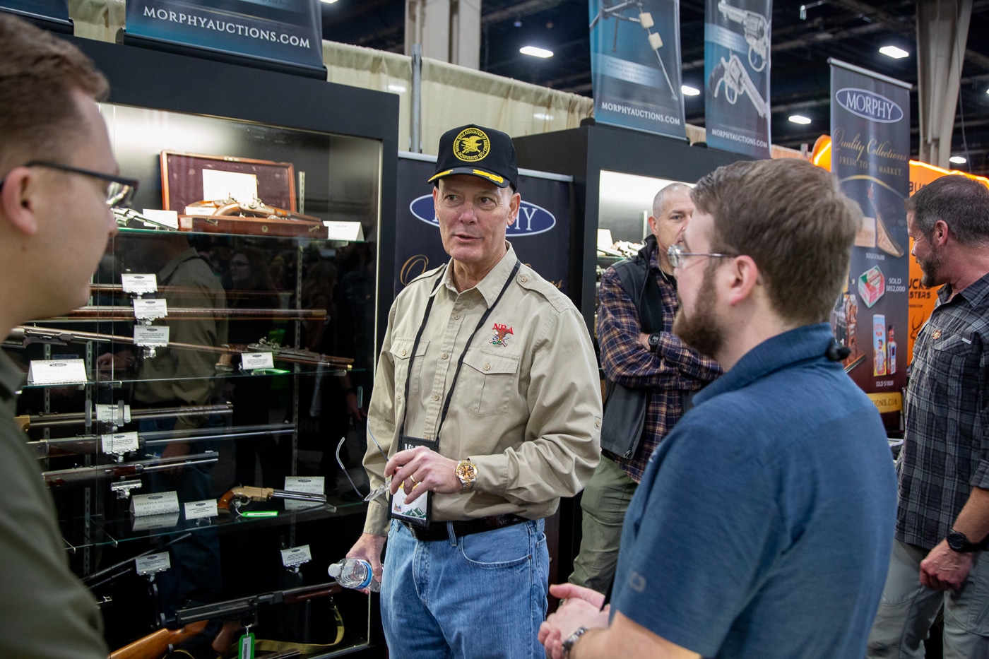 A photo of Doug Hamlin interacting with several people at a busy trade show. He is wearing business attire, standing in the middle of a small group, and speaking with engaged attendees. Booth displays and promotional banners are visible in the background, indicating the event’s professional setting. Hamlin appears approachable, smiling and using open gestures as he converses. The scene emphasizes his role in connecting with both NRA members and industry representatives. It conveys leadership, networking, and accessibility within a public event context.