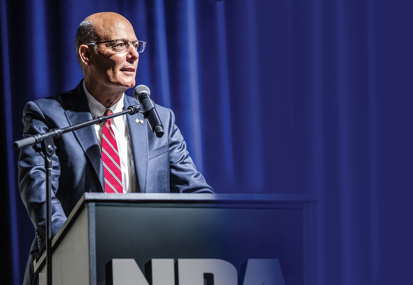 A photo of Doug Hamlin standing behind a podium while giving a speech at a formal event. He is dressed in a suit and tie, speaking into a microphone as attendees listen. The image captures him mid-sentence, with a confident expression that reflects his leadership role as CEO and Executive Vice President of the National Rifle Association. The background shows event signage, suggesting this is an official NRA or firearms-related function. Hamlin’s body language conveys authority and engagement, underscoring his focus on membership trust and organizational accountability. The setting emphasizes his position as a public representative of the NRA.