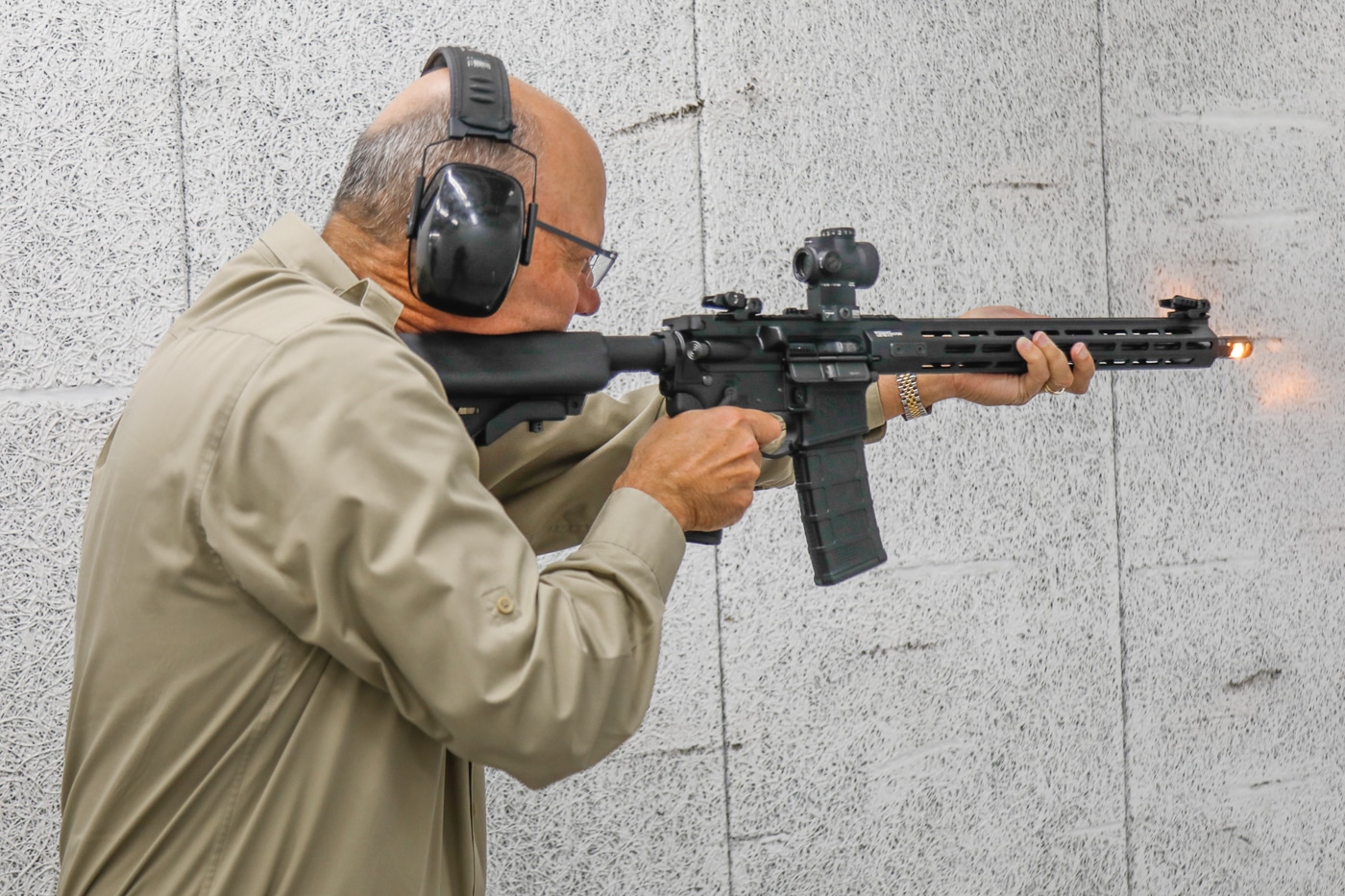A photo of Doug Hamlin on a shooting range, aiming downrange with an AR-style rifle. He is wearing safety glasses and ear protection, dressed casually for range use. The rifle is shouldered properly, with Hamlin maintaining a steady stance as he prepares to fire. The outdoor setting includes targets in the distance and range markers. His posture shows familiarity and experience with the platform. The image underscores his practical use of modern sporting rifles, linking his advocacy role with personal involvement in shooting sports.