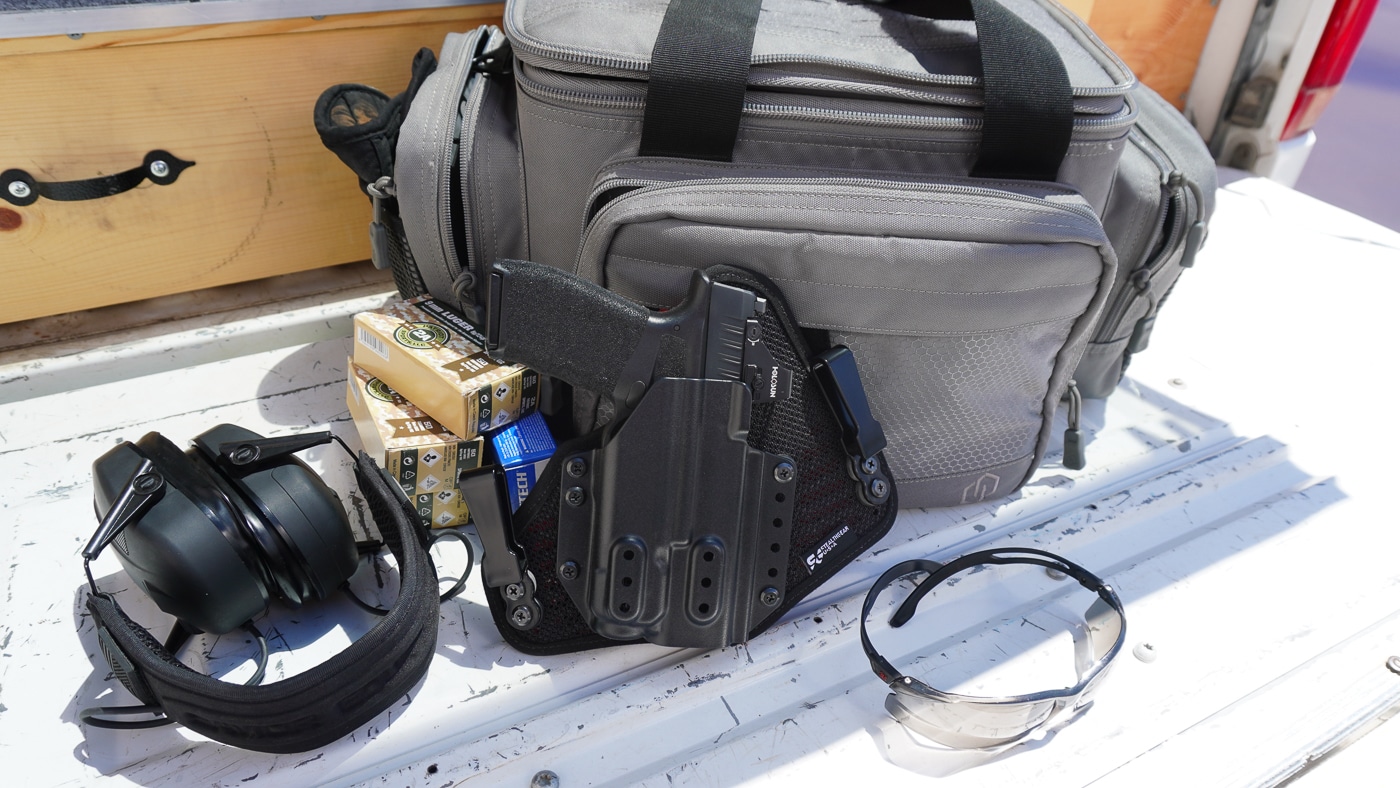 Photograph of a Springfield Hellcat Pro pistol seated in a StealthGearUSA Ventcore 2.0 IWB Mini holster, arranged on a flat surface with range gear. Items like extra magazines, eye and ear protection, and ammunition are visible around the holster. The handgun has a mounted Streamlight TLR-7A weapon light attached, showing compatibility with light-bearing pistols. The Kydex shell and ventilated backing of the holster are clearly seen. The setup looks like a practical loadout for a full day of live-fire training. The arrangement emphasizes concealed carry readiness and gear quality.