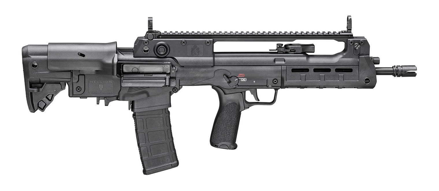 Three-quarter view of a Springfield Hellion bullpup rifle laid on a subdued surface next to a small pack and compact sling, presented to illustrate a practical bug-out gun setup. The picture shows the Hellion’s rear-mounted action and shortened overall length while retaining the full-length barrel inside the compact chassis, which makes it relevant for articles about Springfield Armory bug out guns and vehicle-defense rifles. Ambidextrous controls, the charging handle area, and the handguard are visible, suggesting ease of use for both right- and left-handed carriers without showing handling. The finish and hardware detail are clear, emphasizing a field-ready, durable appearance rather than decorative features. Background items like a folded map or seatback organizer are softly out of focus to imply storage and rapid deployment options. No operational or firing content is depicted; the image is strictly editorial and useful for accessibility and SEO-driven gear roundups.