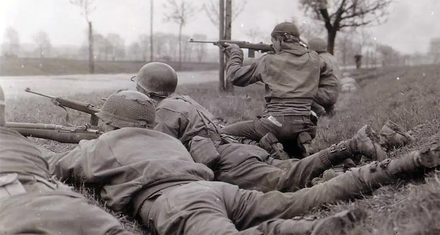 American serviceman wielding M1 Carbine rifle during final stages of World War II combat in Germany April 1945, showing historical military application of shortened barrel carbine design. The lightweight M1 Carbine being used in this wartime photograph chambered .30 Carbine ammunition and weighed significantly less than standard infantry rifles issued to frontline troops. Historical combat image demonstrates the carbine's role as weapon for support personnel, vehicle operators, artillery crews, and officers who needed more firepower than handguns but found full-length rifles impractical. M1 Carbine shown in action represents one of most successful American military carbine designs, with production exceeding six million units during the war. The compact rifle visible in this April 1945 photograph offered soldiers improved maneuverability in urban combat and confined spaces typical of late-war European fighting. Authentic World War II image shows the shortened rifle design that revolutionized military small arms by providing effective firepower in highly portable package suitable for non-frontline troops and close-quarters battle situations.