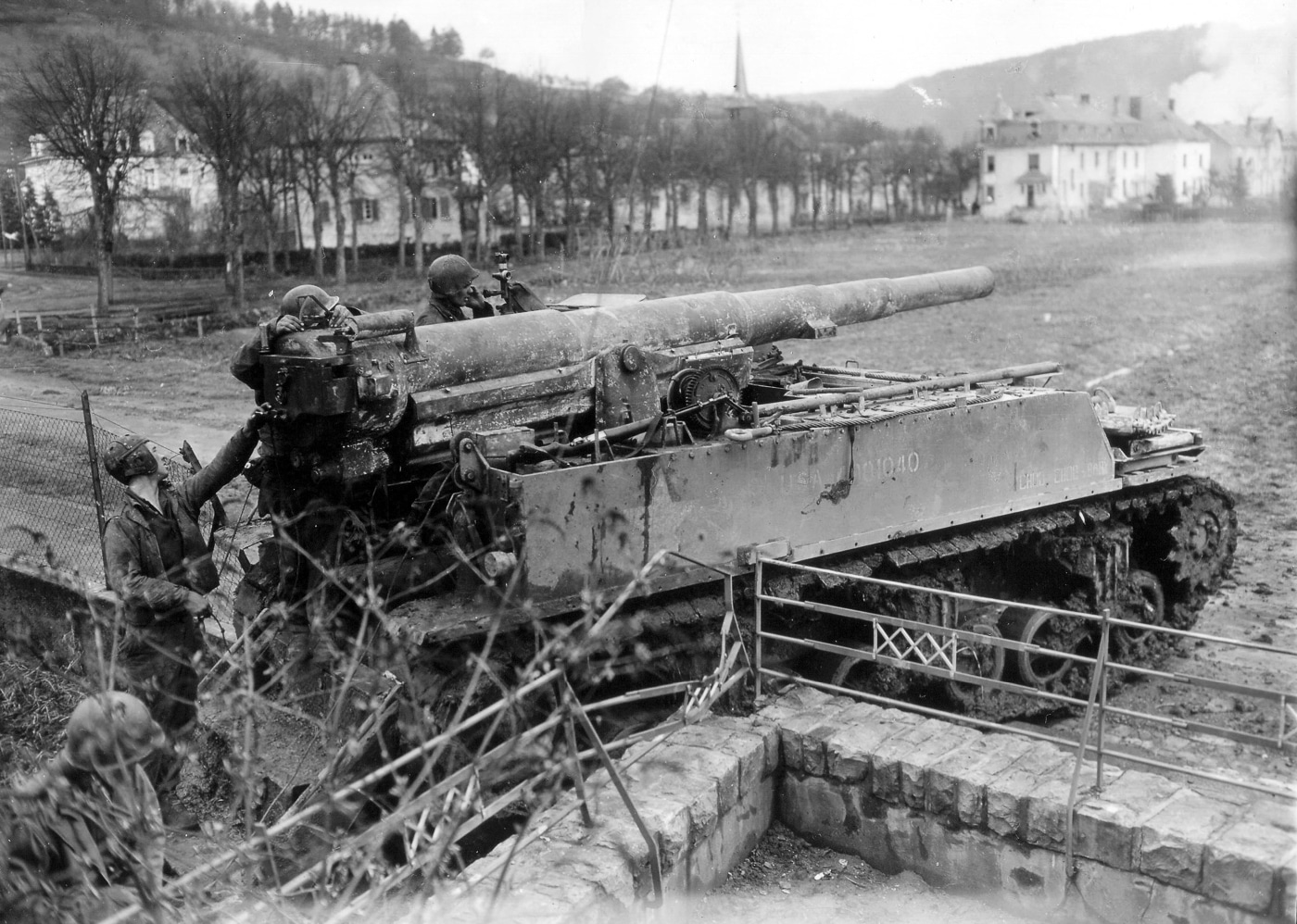 An M12 Gun Motor Carriage is shown firing its 155mm cannon at Siegfried Line bunkers near Echternach, Germany, in 1945. The vehicle is dug in with its rear spade deployed to absorb recoil, stabilizing the chassis during firing. The long barrel is elevated, and smoke from the discharge suggests the weapon is mid-action. These heavy self-propelled guns were among the few capable of penetrating six feet of reinforced concrete with multiple hits. The open-topped design exposes the gun crew even as they deliver devastating firepower. Ammunition was limited to ten rounds onboard, requiring resupply from companion M30 cargo carriers. This image captures the raw power and tactical importance of the M12 during the campaign against the German Westwall defenses.