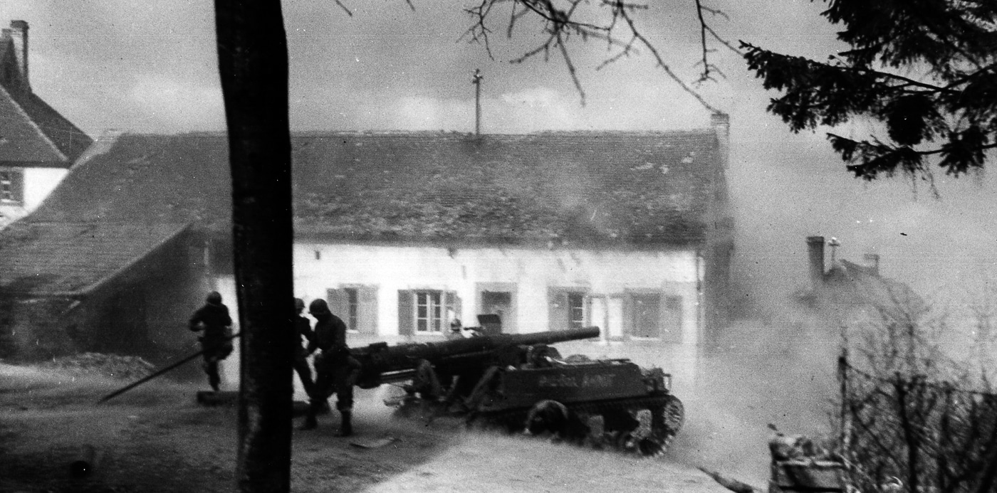 An M12 Gun Motor Carriage is captured firing near Seelbach, Germany, in 1945 while supporting the U.S. 45th Infantry Division. The vehicle mounts a 155mm gun on a modified M3 tank chassis, its barrel extended and recoiling as a shell is discharged. The rear spade is deployed in the ground to stabilize the vehicle against the force of firing. The open-topped fighting compartment exposes the crew, who operated under dangerous conditions during combat missions. The M12 carried only ten rounds of ammunition, requiring constant resupply by M30 cargo carriers. Despite these limitations, its firepower was unmatched, able to smash bunkers and pillboxes along the German defensive line. This image highlights how mobile artillery worked in direct coordination with infantry divisions in the final year of the war.