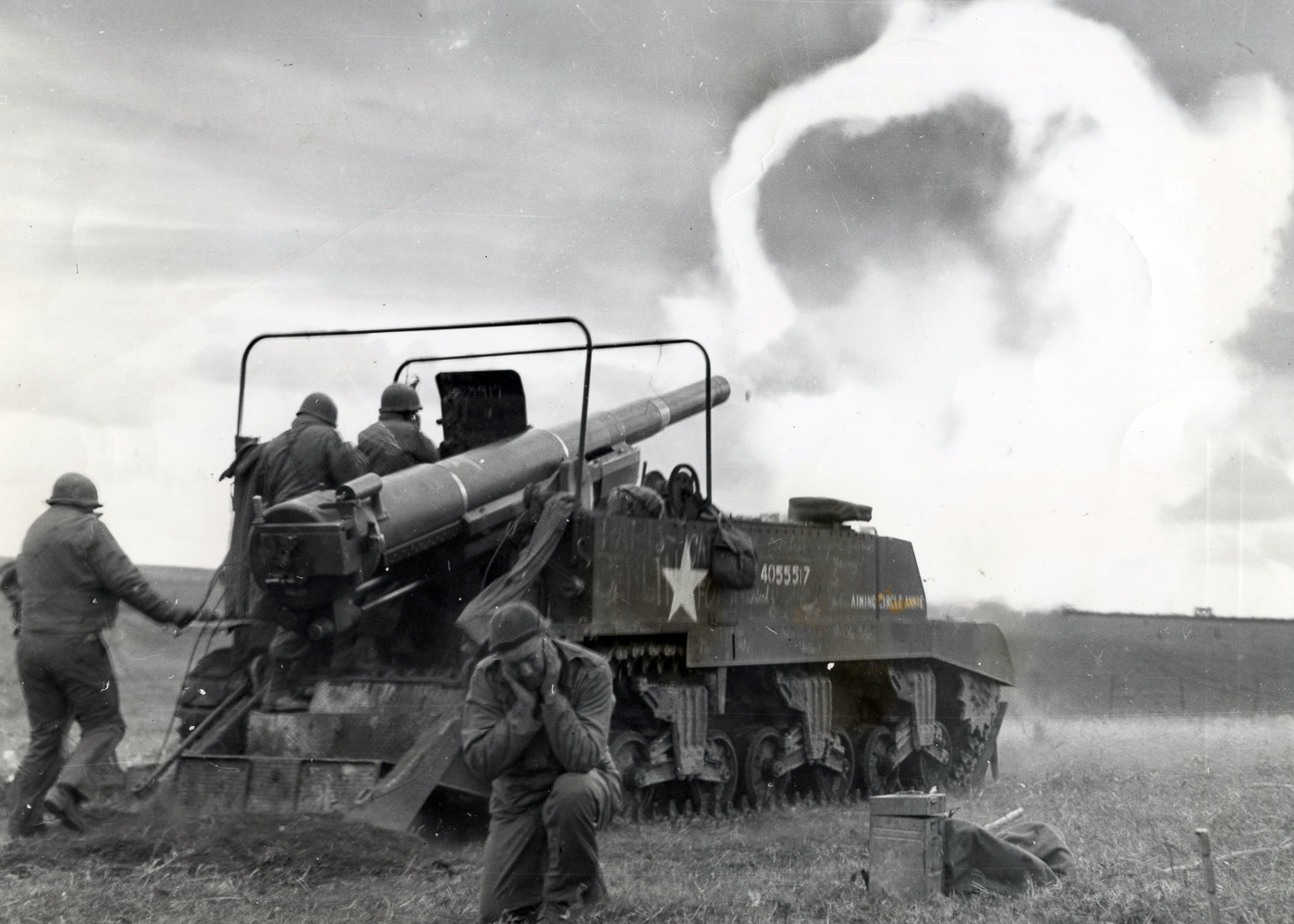 An American M12 Gun Motor Carriage is captured in the middle of firing its 155mm cannon during combat in France in World War II. The long gun barrel is elevated, and the blast produces a visible recoil effect from the open-topped vehicle. The crew operated from an exposed fighting compartment at the rear, making them vulnerable even while delivering heavy fire. The rear spade of the vehicle, dug into the ground, stabilizes the chassis against the enormous recoil. The M12 was often used for direct fire missions against bunkers and fortified positions, especially during the advance across France. This scene demonstrates the balance of raw firepower and limited protection that defined the M12’s role. The photograph highlights the intensity of mobile artillery in European combat operations.