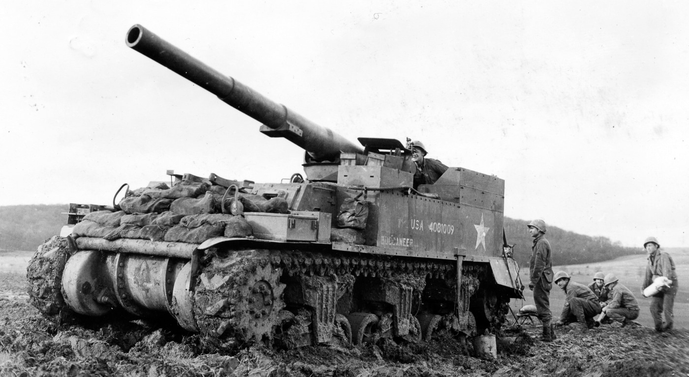 An American M12 Gun Motor Carriage is parked in Germany in the spring of 1945, reinforced with sandbags stacked across the front and sides. The vehicle carries a 155mm gun mounted on the M3 tank chassis, making it a powerful but vulnerable self-propelled weapon. The open-top crew area is visible, showing why soldiers added improvised protection against shrapnel and small arms fire. The large spade at the rear, designed to dig into the ground and absorb recoil when firing, can also be seen. This image highlights the field modifications U.S. forces often made to keep the M12 in service during heavy fighting. The use of sandbags was a practical solution, even if it added weight and slowed movement. The photo documents how artillery crews adapted their equipment in the last months of World War II.