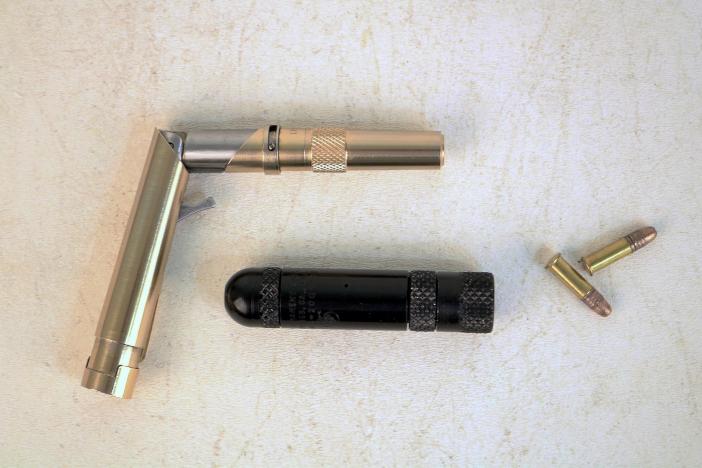 The image displays a MAC Stinger pistol, a Braverman Stinger pen gun, and several .22 LR cartridges. The MAC Stinger, positioned as a small cylindrical firearm, is only about 3.25 inches long, while the Braverman has a folding design resembling a pen when collapsed. The inclusion of the .22 LR rounds emphasizes the small caliber both firearms were designed to fire. The scale makes clear how tiny these weapons are, especially compared to conventional pistols. Both guns were classified by the BATFE as “Any Other Weapons” due to their nontraditional construction. While compact and easily concealable, they were considered unsafe and ineffective for practical use. The photo provides a clear look at these rare Cold War-era novelty firearms and their ammunition.