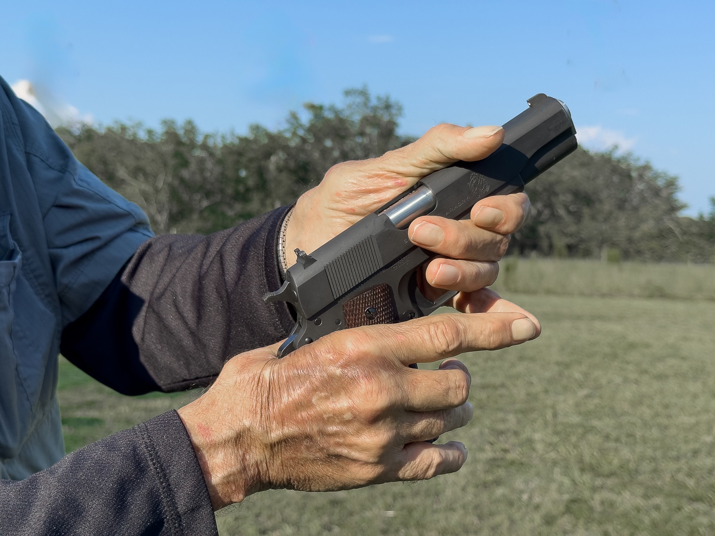 Massad Ayoob stands in front of a training audience while holding a 1911 pistol, demonstrating a common mistake shooters make when learning to operate the firearm. His focus is on safety and proper handling of the gun. The photo captures his hand placement on the pistol, showing how small errors can affect function. Ayoob’s teaching emphasizes avoiding habits that cause malfunctions. The 1911 design requires specific techniques to use effectively. The image shows him correcting errors that are often passed down in training. This visual highlights both instruction and firearm awareness.