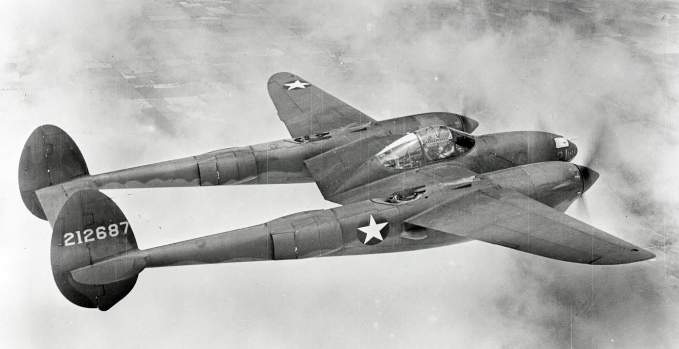 Black-and-white photograph of a Lockheed P-38 Lightning, highlighting its distinctive twin-boom tail and central nacelle cockpit design. The plane was unlike any other Allied fighter of the era, built for long-range missions and capable of carrying heavy firepower. Four .50 caliber machine guns and a 20mm cannon were mounted in the nose, giving it concentrated striking power. The twin-engine layout provided both speed and endurance, allowing it to escort bombers and intercept enemy aircraft far from base. Pilots praised its ruggedness, even though the engines were sometimes vulnerable. Its unique silhouette made it easy to identify in the air. The P-38 became famous for its role in Operation Vengeance and other Pacific missions.