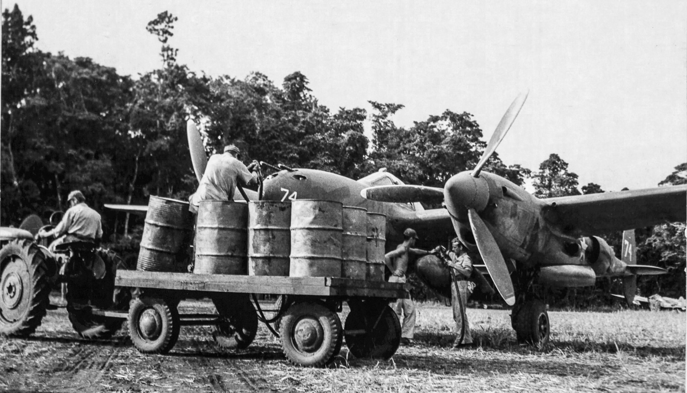 Historical image from 1943 showing a P-38 Lightning fighter on an airstrip in New Guinea while crews prepare it for another mission. The plane’s large wingspan and dual tail booms dominate the frame, while maintenance staff work quickly on refueling. New Guinea was a vital hub for Allied forces, providing airfields from which fighters could strike deep into Japanese-controlled areas. The P-38’s extended range allowed missions that other aircraft could not attempt. Ground crews, often working in challenging jungle conditions, kept these aircraft flying daily. This image illustrates the constant preparation behind the high-profile combat missions of the Pacific war.