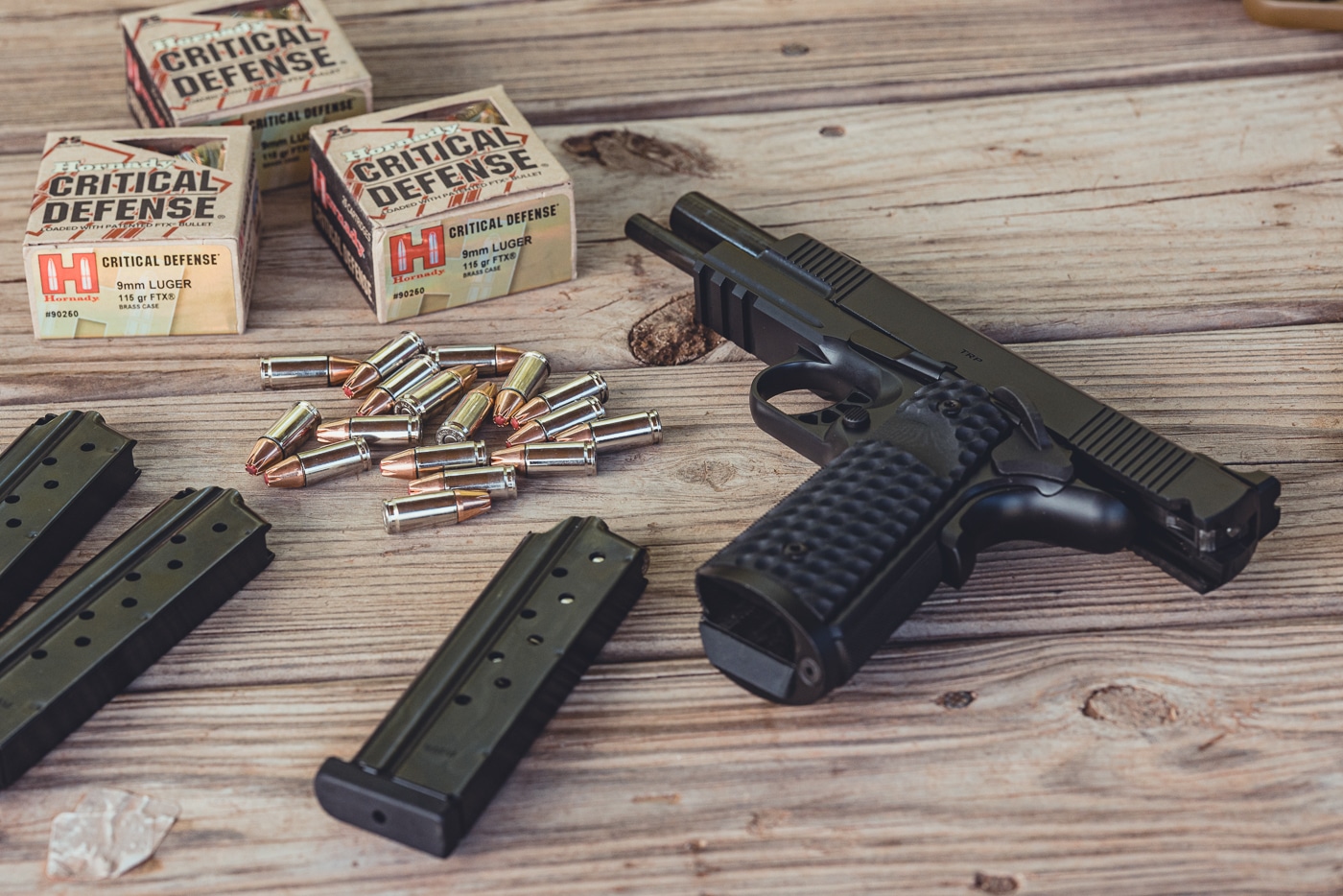 A Springfield Armory 1911 TRP pistol is displayed on a wooden shooting bench next to a box of Hornady Critical Defense ammunition. The handgun is configured with its traditional iron sights, showcasing Springfield’s precise machining and rugged design. The pistol features G10 grips, front strap checkering, and a skeletonized trigger that reflects the TRP series’ performance-oriented build. The Hornady Critical Defense ammo highlights the pistol’s use in self-defense applications, pairing a classic firearm design with modern ammunition technology. The wood bench surface provides a natural backdrop, emphasizing a real-world range or training environment. This image captures the practical and reliable side of the 1911 platform.