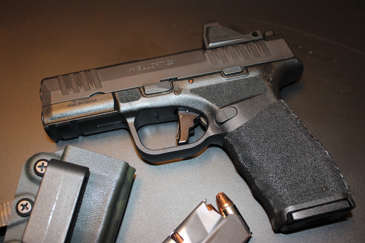 Front three-quarter photo of a Springfield Armory Hellcat Pro pistol placed on a flat, neutral surface, shown as a compact option for bug-out and concealed carry purposes. The black polymer frame and textured grip are visible, along with the optics-ready slide that can accept a small red dot sight. The magazine well is flush with the frame and a spare magazine sits nearby, indicating higher capacity than many micro-compact pistols. The trigger face and slide serrations are clearly shown, and the pistol’s compact overall length and low profile are emphasized to illustrate portability. This image is suitable for articles about Springfield Armory bug out guns, compact self-defense pistols, and emergency carry gear. No people are present and the background is unobtrusive to keep the focus on the Hellcat Pro’s features.