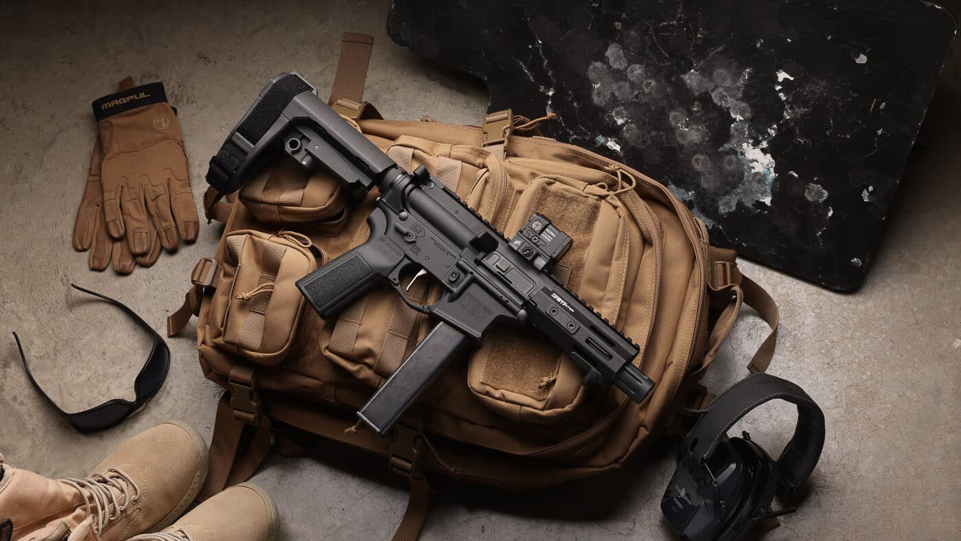 Photo shows a Springfield Armory SAINT 9mm with a 5.5” barrel placed on top of a tactical backpack alongside common bugout items such as a medkit, water bottle, and multitool. The firearm is displayed with the pistol brace collapsed to emphasize its compact dimensions for transport and rapid deployment. A small red-dot optic is visible on the top rail, promoting fast target acquisition during short-range engagements typical in urban defense and CQB scenarios. The image highlights the SAINT Victor 5.5” 9mm’s balance of portability and defensive firepower, ideal for a 9mm carbine configuration in a grab-and-go kit. Viewers can see the slim handguard and reduced overall length that reduce snag points when moving through vehicles or tight spaces. Keywords included: SAINT Victor 5.5” 9mm, Springfield Armory SAINT, bugout gear, compact AR, 9mm carbine.