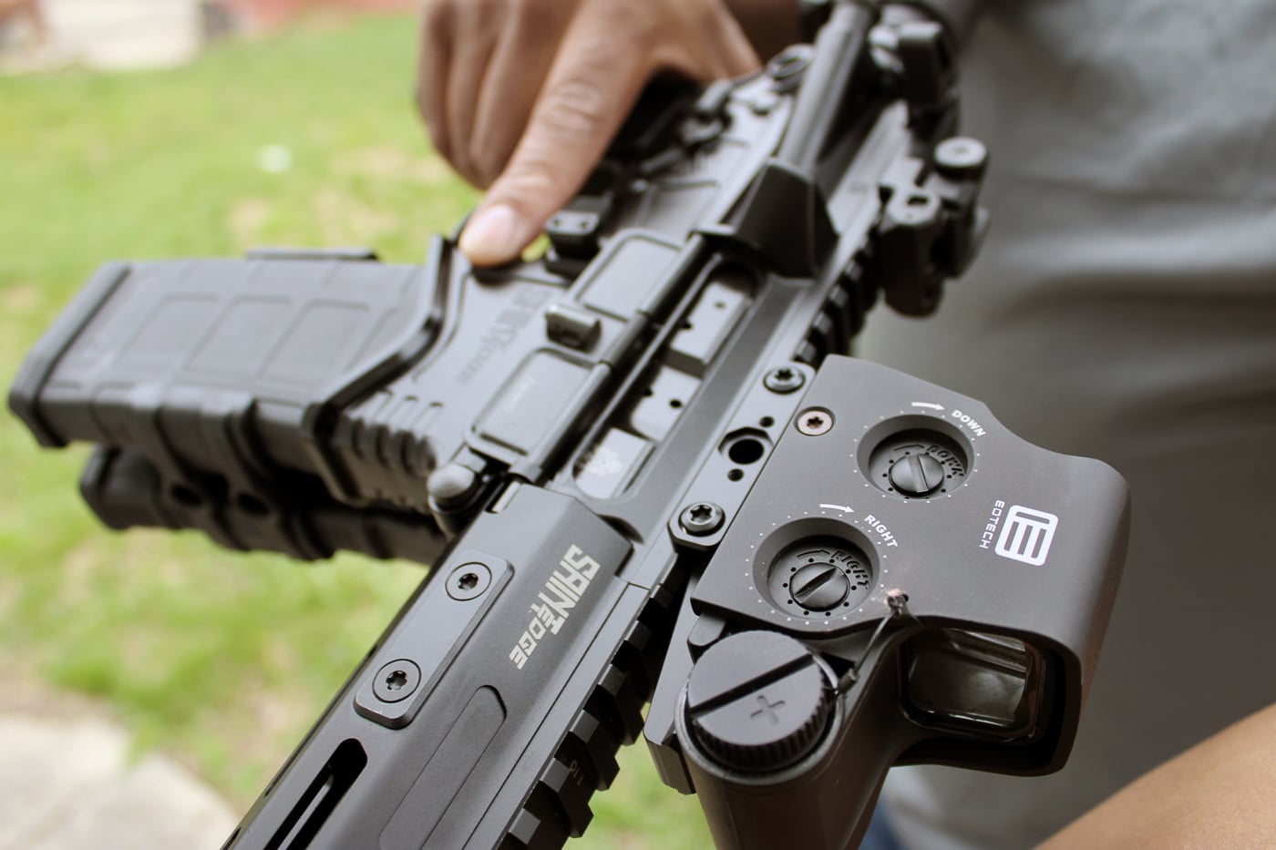 Close-up image of a Springfield Armory SAINT Edge rifle chambered in 5.56, photographed horizontally on a dark surface with an EOTech EXPS red dot sight mounted on the top rail. The black AR-style lower and free-floating M-LOK handguard are visible, along with the short muzzle device and collapsible stock. The EOTech EXPS optic sits low to the bore for quick sight picture and is clearly labeled on the housing. This image highlights the rifle’s compact profile and accessory-ready design, suitable for discussions of Springfield Armory bug out guns and home defense rifles. The lighting emphasizes texture on the grip and rail, suggesting durability and real-world use. No people are visible in the photo, and the background is intentionally unobtrusive to focus attention on the SAINT Edge and optic.