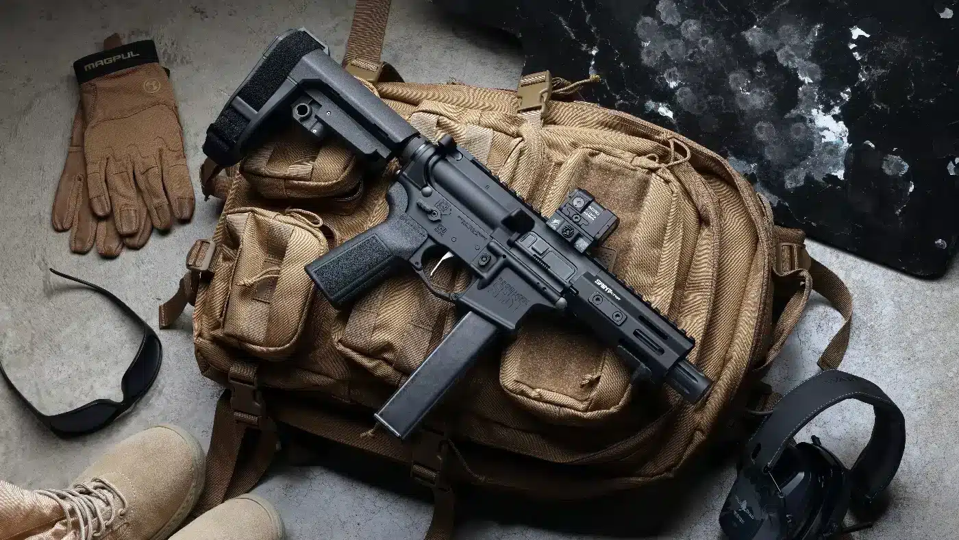 Image of a Springfield Armory SAINT 9mm carbine with a 5.5-inch barrel photographed on a neutral surface, displayed as a compact bug-out gun option. The short barrel and compact collapsible stock are visible, emphasizing portability and easy storage in a pack or vehicle. The lower and upper receivers show standard AR-style controls, and the M-LOK handguard provides mounting points for lights or sling hardware. The magazine appears seated and the chamber area is closed, showing a non-operational gear shot rather than a handling demonstration. This photo is useful for content about Springfield Armory bug out guns, compact 9mm carbines, and emergency preparedness gear. Lighting is even to show texture and finish without distracting reflections.