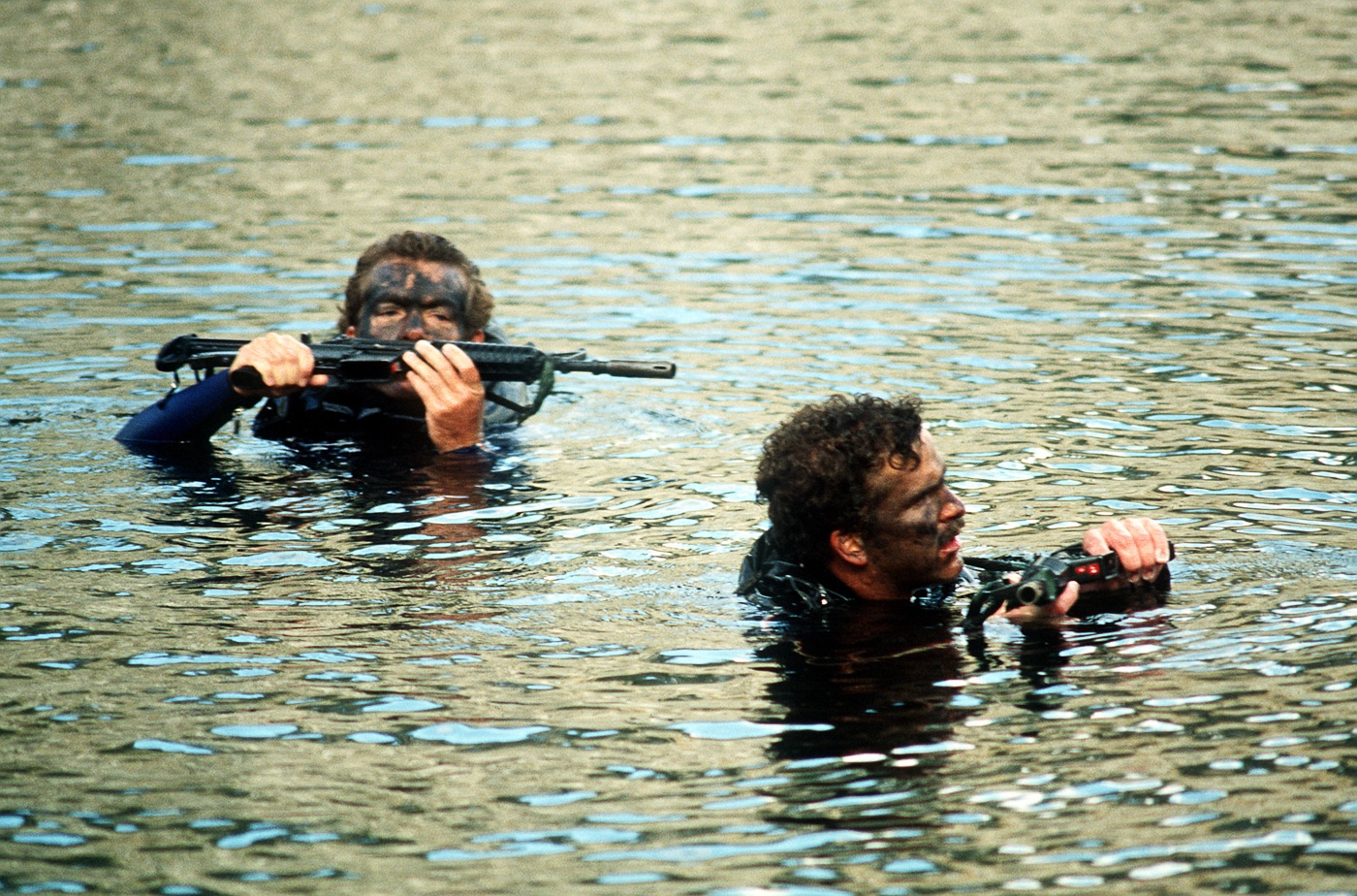 U.S. Air Force combat controllers are pictured in a 1970s–1980s era training scenario, wading through a river while armed with XM177 rifles. The carbines are visibly compact, featuring moderator flash hiders, collapsible stocks, and standard-issue 20-round magazines. Each man holds his rifle above shoulder height to keep it dry while crossing. Their field gear includes helmets, web belts, and radios, marking them as specialized operators rather than regular infantry. The scene highlights the challenges of special operations mobility in waterborne environments. XM177, CAR-15, Colt Commando, combat control team, river navigation, special operations carbine.