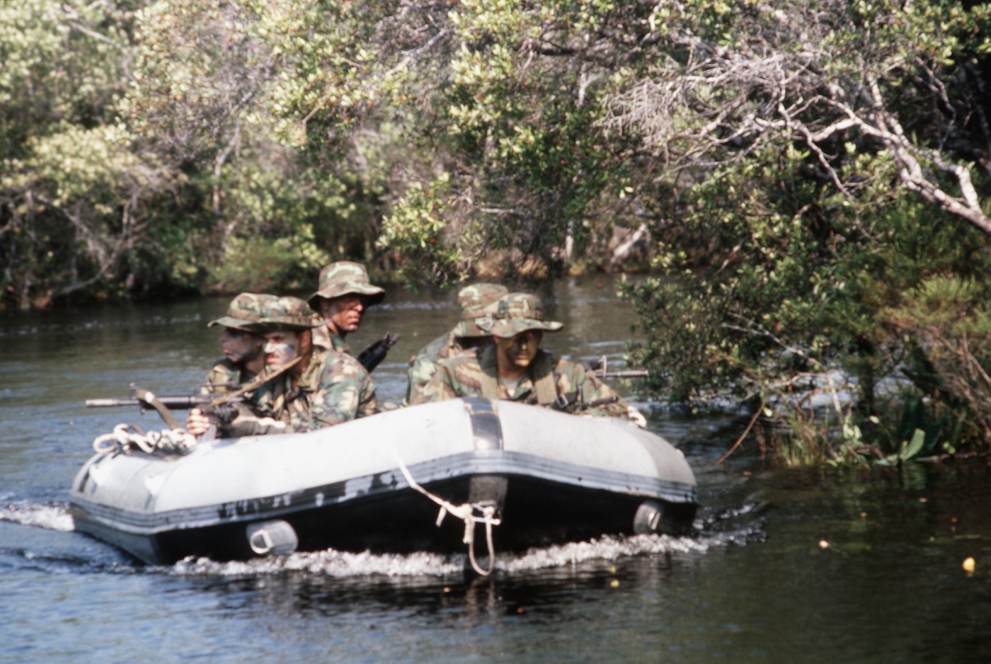 The photo depicts a U.S. Air Force combat control team seated in an inflatable boat during a training exercise, each man armed with a GAU-5 variant of the XM177 carbine. The carbines are identifiable by their short barrels with moderator muzzle devices, collapsible aluminum stocks, and inserted 20-round 5.56mm magazines. Team members wear field uniforms, helmets, and tactical web gear, with radios and equipment visible inside the boat. The inflatable boat is in calm water, suggesting a river or coastal training area. The compact rifles are held at the ready, emphasizing their role in rapid-action maritime operations.  GAU-5, XM177, CAR-15 Commando, USAF combat control, inflatable boat training, 5.56mm carbine.