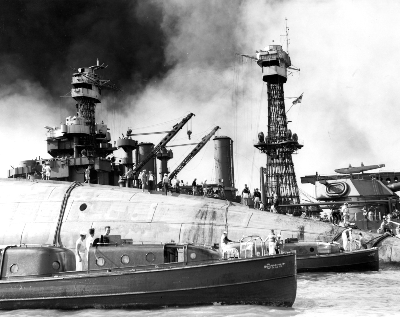 The photograph shows rescue crews working on the overturned hull of USS Oklahoma (BB-37) at Pearl Harbor on December 7, 1941. The battleship lies completely capsized, with her starboard bilge keel visible above the waterline. Teams of sailors and workers stand on the exposed hull, cutting into the steel in an attempt to reach trapped crew members inside. The Oklahoma had been hit by multiple torpedoes during the Japanese attack, causing her to roll over and sink quickly. Hundreds of sailors were killed, though some were rescued through holes cut into the hull. The image highlights both the scale of destruction at Pearl Harbor and the desperate rescue efforts carried out in the immediate aftermath.