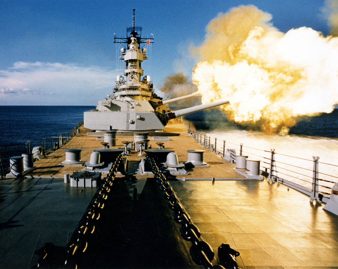 The photo shows the USS Wisconsin (BB-64) in action, firing her 16-inch main battery guns during a live-fire exercise. Massive bursts of flame and smoke erupt from the barrels as the shells leave the gun turrets, producing shockwaves across the ocean surface. The ship’s deck and superstructure are partially obscured by the smoke cloud, emphasizing the sheer force of the blast. Each shell from these guns could weigh around 2,700 pounds and reach targets more than 20 miles away. The scene highlights the raw destructive capability that made battleships central to naval warfare during World War II and into the Cold War. The bright muzzle flashes contrast against the gray ship and ocean, underlining the violent energy of the firing sequence. This dramatic image captures the power and spectacle of a battleship’s gunnery, even as such weapons became increasingly outdated in modern combat.