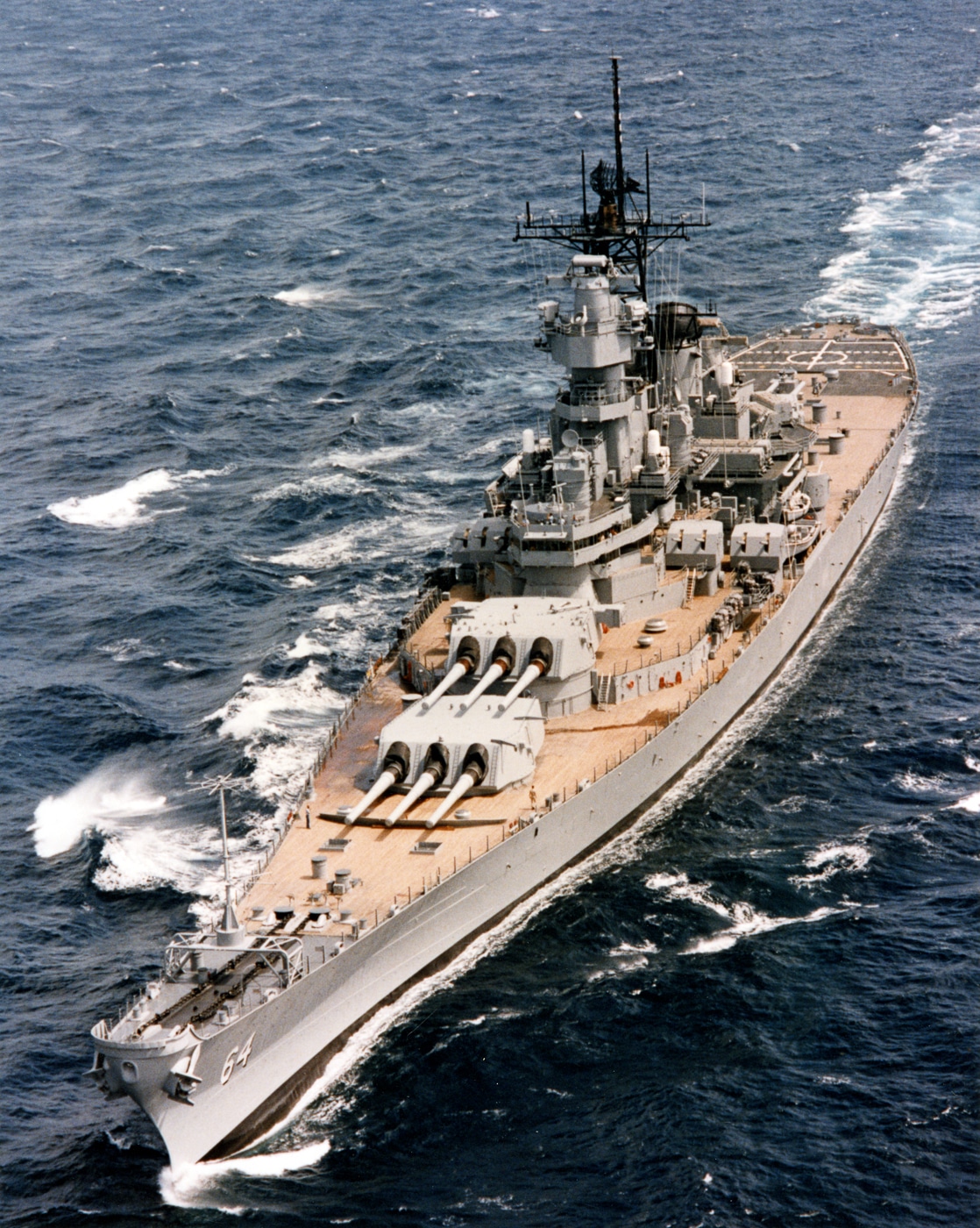 The photo shows the USS Wisconsin (BB-64), one of the U.S. Navy’s Iowa-class battleships, sailing at sea in 1989. The massive warship cuts through the water with her long gray hull and towering superstructure. Visible on deck are the ship’s iconic 16-inch gun turrets, arranged in triple mounts, alongside modern missile systems that were added during Cold War refits. Radar masts and communication antennas rise above the bridge, reflecting her updated role in missile-era warfare. A white wake trails behind the ship as she moves at speed across open water. Despite being designed in the 1940s, her silhouette looks imposing and combat-ready. This image captures one of the last active deployments of a U.S. battleship before the class was permanently retired.