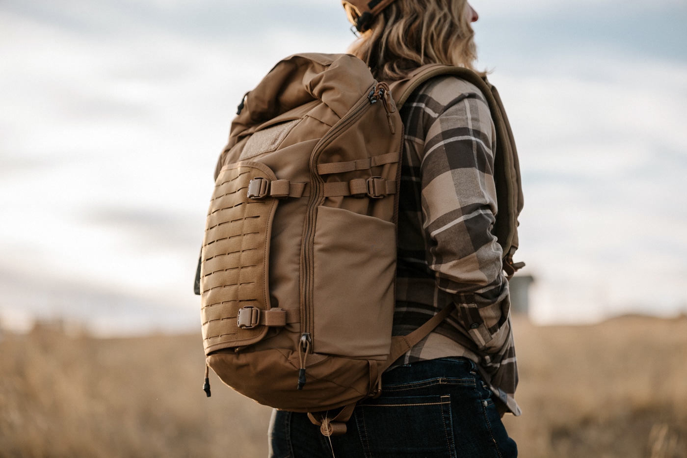 Photo of a woman carrying the Vertx Siege 25L Tactical Backpack during an in-depth review process. The pack is worn on her back, showing adjustable padded shoulder straps and a ventilated mesh back panel for comfort. The sleek black nylon exterior with MOLLE webbing provides tactical durability without looking overly military. Side compartments and the expandable front pocket are visible, designed to hold water bottles, gear, or larger items. The waist and chest straps help distribute weight evenly for long periods of wear. The review emphasizes ergonomic fit for both men and women in everyday and field use. The photo illustrates practical carry testing as part of the product evaluation.