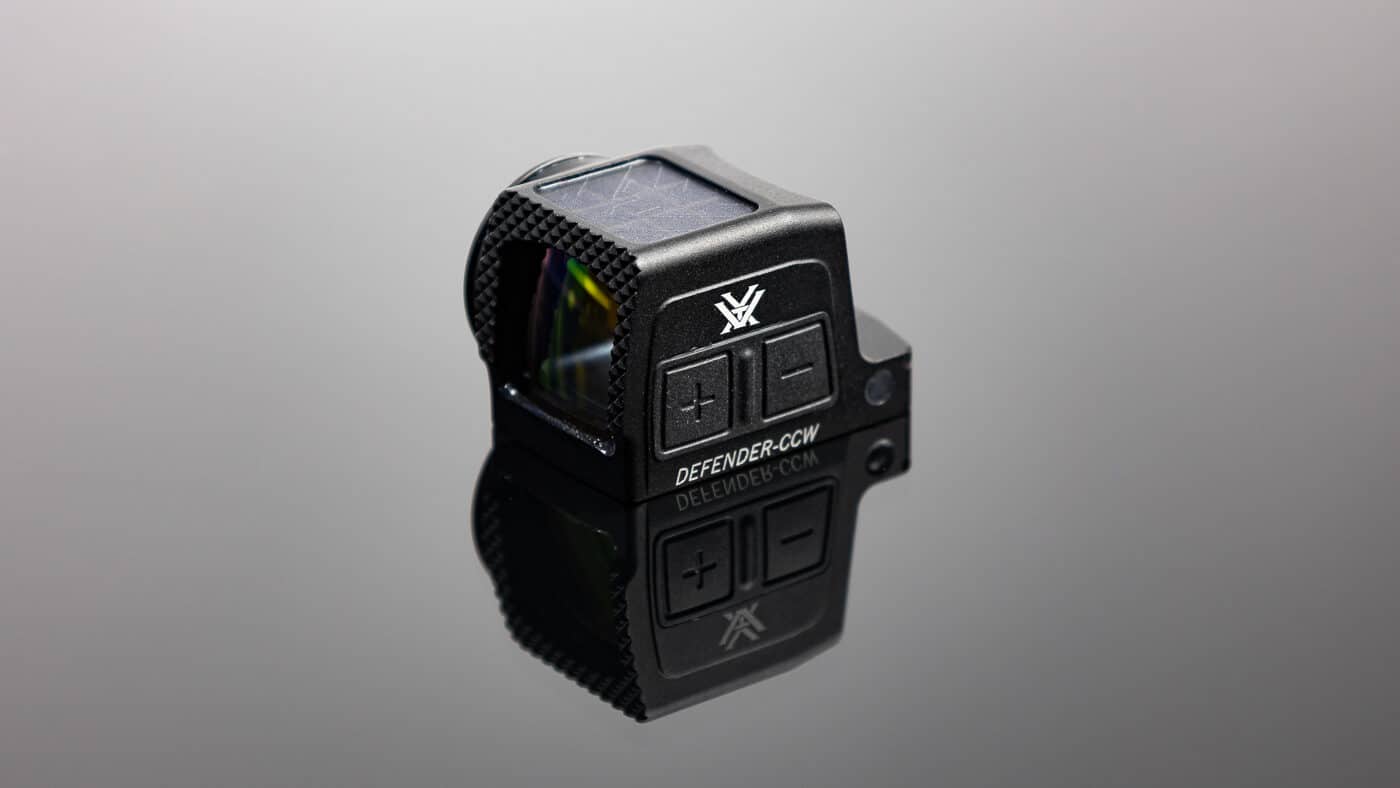 Detailed image of the Vortex Defender-CCW Enclosed red dot sight mounted on a compact handgun slide. The optic has a short housing measuring about 1.6 inches long and weighing just over an ounce with its battery installed. Its black anodized 7075 aluminum body features Fast-Rack texturing at the front for one-handed slide manipulations. The enclosed emitter design keeps the LED protected from sweat, lint, dust, and rain, ensuring reliability for concealed carry. On the side of the optic, the battery compartment is visible, allowing quick battery changes without dismounting. The large viewing window offers a clear sight picture for faster target acquisition. The sight sits low on the slide to maintain compatibility with standard-height pistol sights.